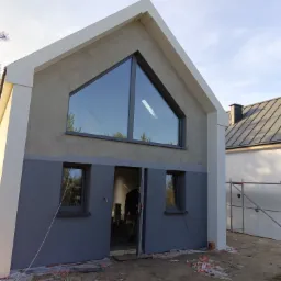 Dwukondygnacyjny budynek w trakcie prac elewacyjnych, z widocznym oknem trójkątnym, częściowo pomalowaną fasadą na szaro i białymi elementami wykończeniowymi, rusztowaniem w tle i rozrzuconymi...