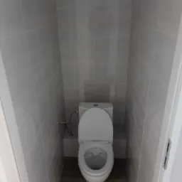 Wąska toaleta w odcieniach szarości, z płytkami imitującymi beton i drewnianą podłogą, widok z perspektywy wejścia.