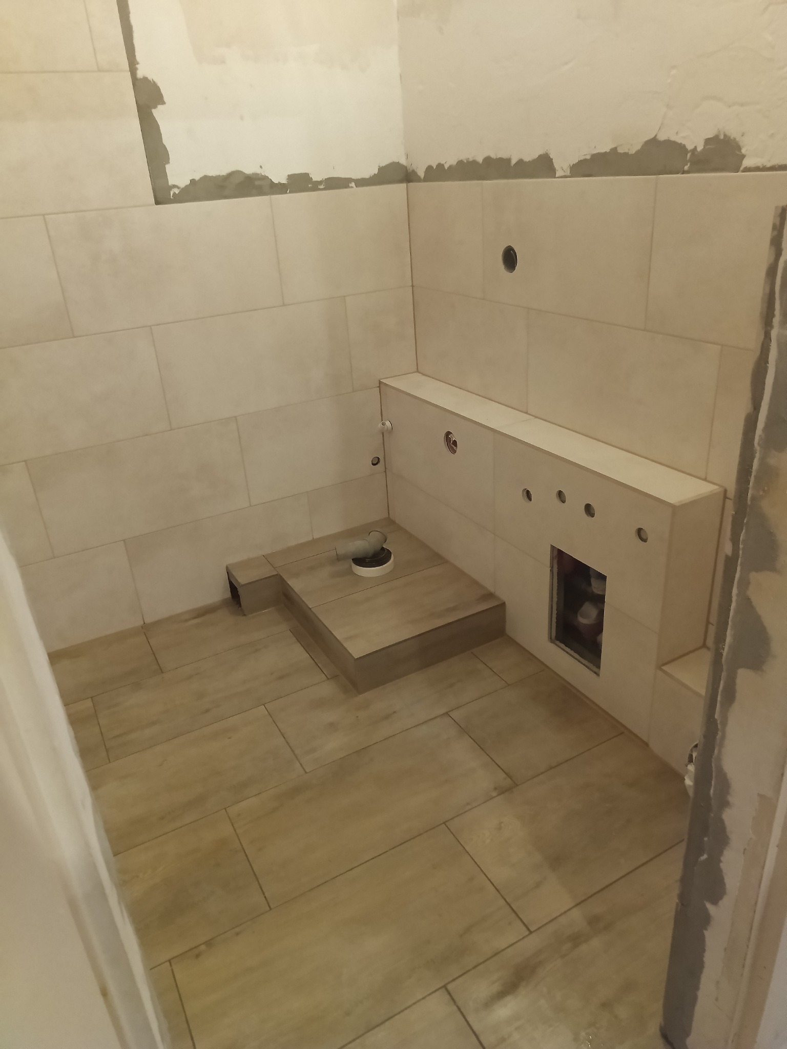 Łazienka w trakcie kładzenia płytek, widoczne płytki na podłodze i ścianach, zabudowa stelaża WC, otwory pod instalacje, fragment niezakończonej ściany z widocznym klejem.