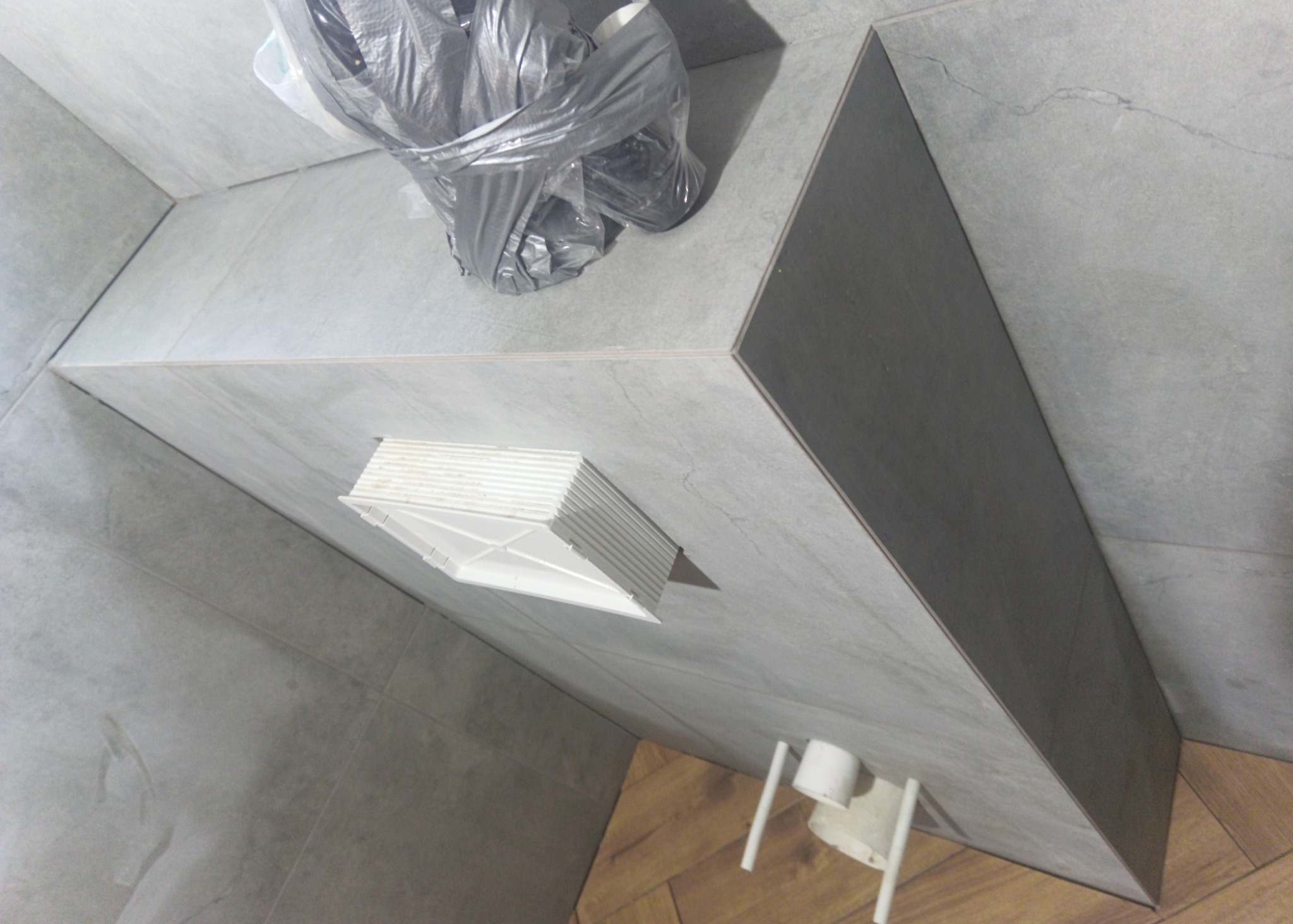 Narożnik łazienki wyłożony szarymi płytkami imitującymi kamień, z widoczną kratką wentylacyjną, workiem na śmieci i fragmentem instalacji toaletowej, na podłodze panele drewnopodobne.