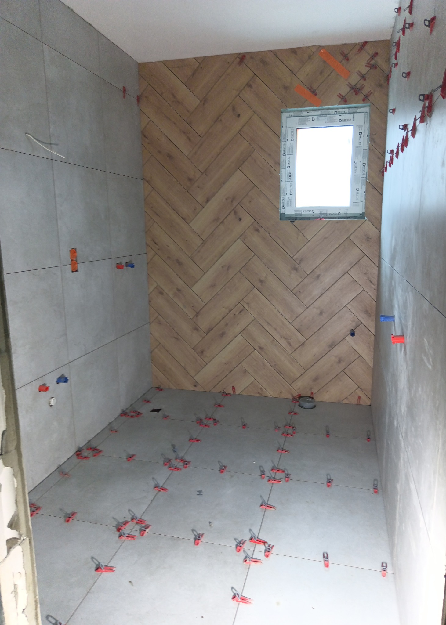Łazienka w trakcie układania płytek: szare płytki na ścianach, drewnopodobna mozaika w jodełkę na jednej ścianie z oknem, czerwone i niebieskie klipsy poziomujące na podłodze i ścianach.