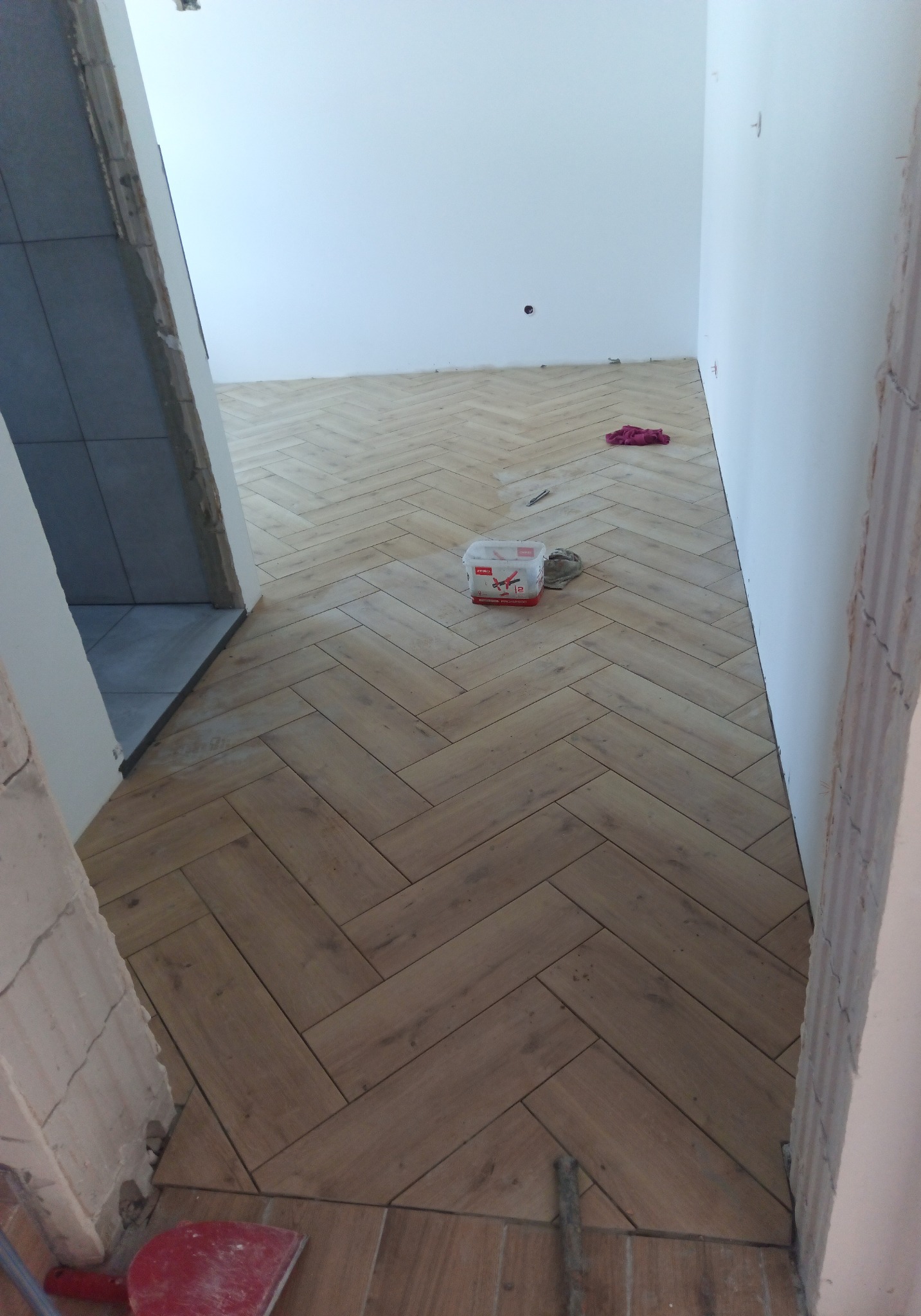 Ułożona podłoga z płytek imitujących drewno w jodełkę, widoczny pojemnik z klejem i czerwona szpachelka przy progu, surowe ściany w trakcie prac wykończeniowych.