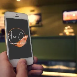 Dłoń trzyma smartfon prezentujący logo 'Hair Care Agnieszka Cieślar' z elementami graficznymi w odcieniach pomarańczy, na rozmytym tle wnętrza lokalu.