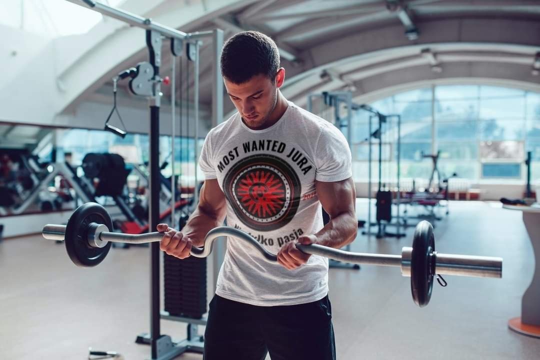 Młody mężczyzna w siłowni, ubrany w białą koszulkę z nadrukiem 'Most Wanted Jura', ćwiczy biceps z użyciem sztangi łamanej.