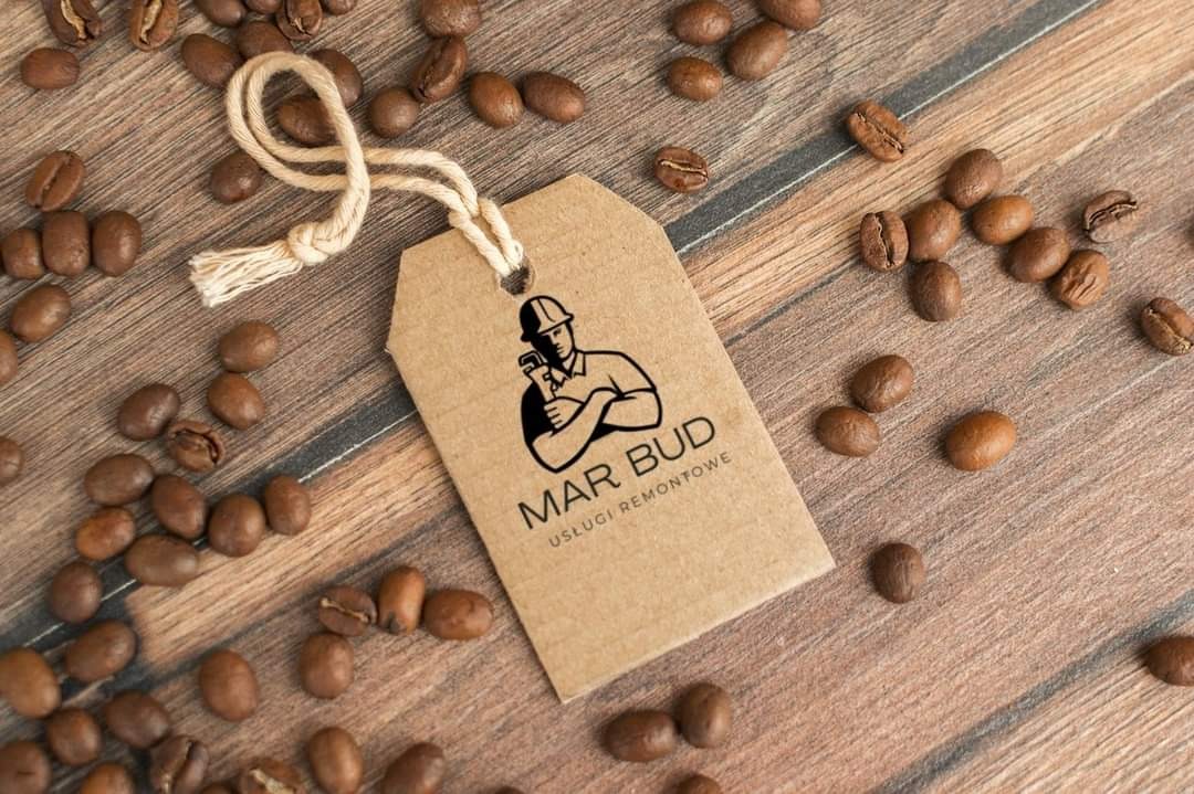 Kraftowa zawieszka z logo 'MAR BUD Usługi Remontowe', przedstawiająca sylwetkę hydraulika w kasku, leży na drewnianym tle otoczona ziarnami kawy.
