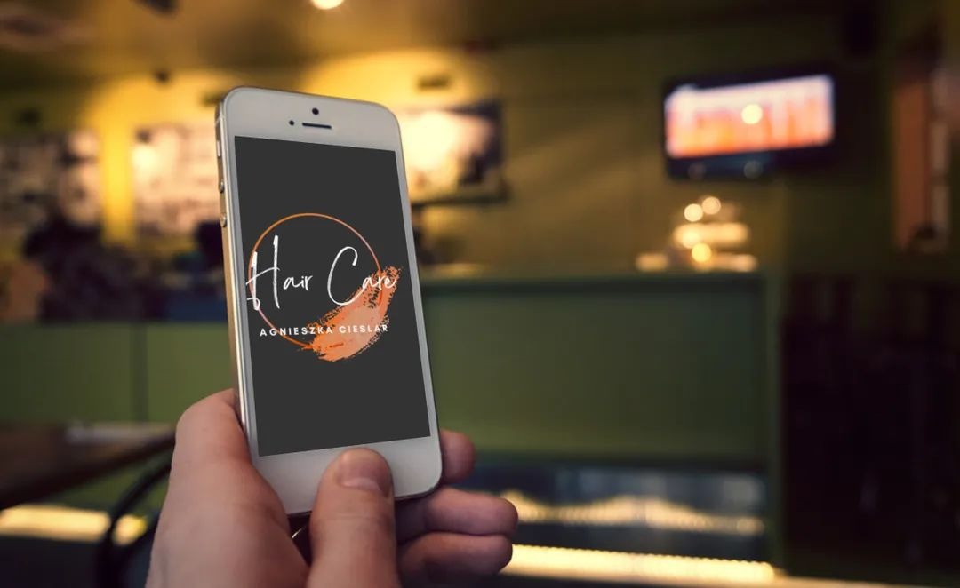 Dłoń trzyma smartfon prezentujący logo 'Hair Care Agnieszka Cieślar' z elementami graficznymi w odcieniach pomarańczy, na rozmytym tle wnętrza lokalu.