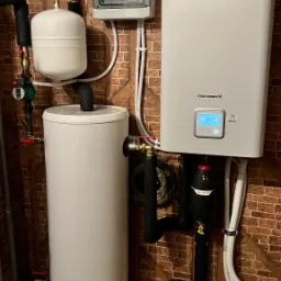 Zestaw grzewczy: Biały zbiornik Galmet, pompa ciepła LG Therma V z wyświetlaczem wskazującym 45 stopni, czarny filtr Fismco, oraz instalacja hydrauliczna na tle ściany z cegły.