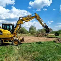 Żółta koparka Komatsu PW98MR przygotowuje teren pod budowę, widoczny wyrównany obszar ziemi i pryzmy usuniętej darni na tle zielonej trawy i błękitnego nieba z chmurami.