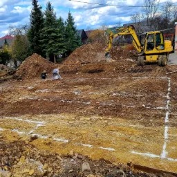 Wytyczony teren pod budowę z widocznymi fundamentami, żółta koparka Komatsu i dwaj pracownicy w tle, ziemia usypana w kopiec, błękitne niebo z chmurami.