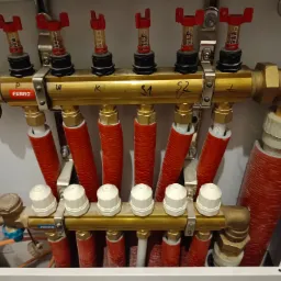 Rozdzielacz instalacji grzewczej marki Ferro, z czerwonymi rurami i przezroczystymi przepływomierzami, zamontowany w szafce, widoczne oznaczenia P, W, K, S1, S2.