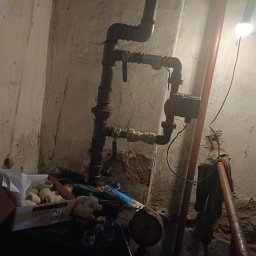Rafał B - Fragment instalacji grzewczej z pompą obiegową, zaworami i rurami w piwnicy z oświetleniem żarówką, widoczne narzędzia i elementy instalacyjne w pudełku.