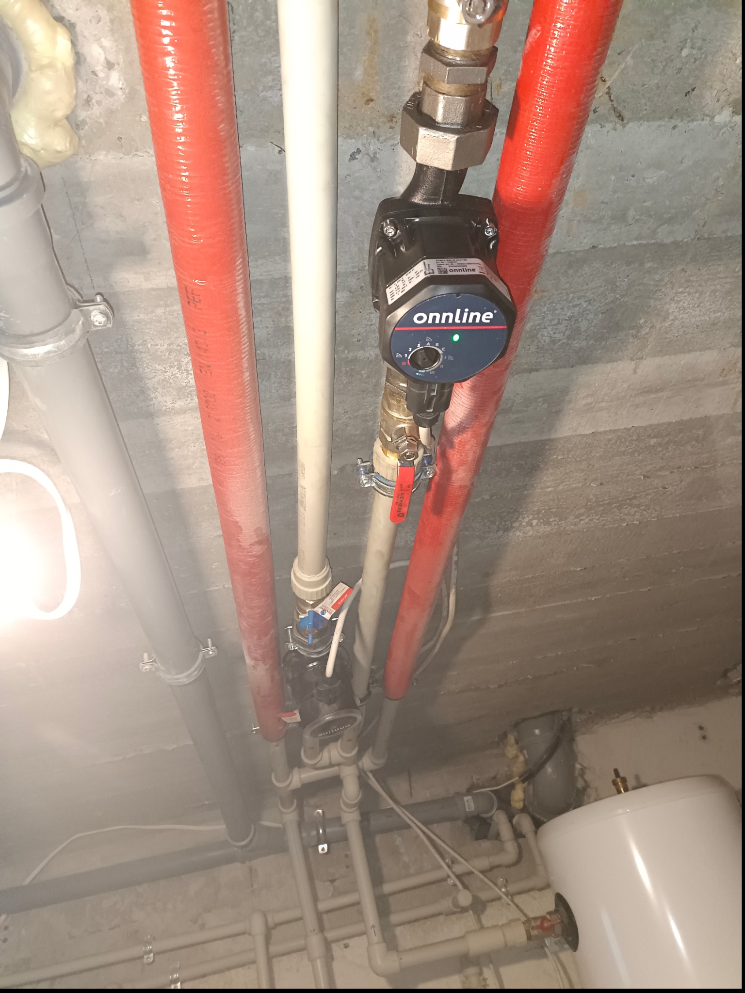 Widok z dołu na instalację hydrauliczną z czerwonymi rurami izolowanymi, pompą obiegową Onnline i bojlerem elektrycznym w szarej piwnicy.