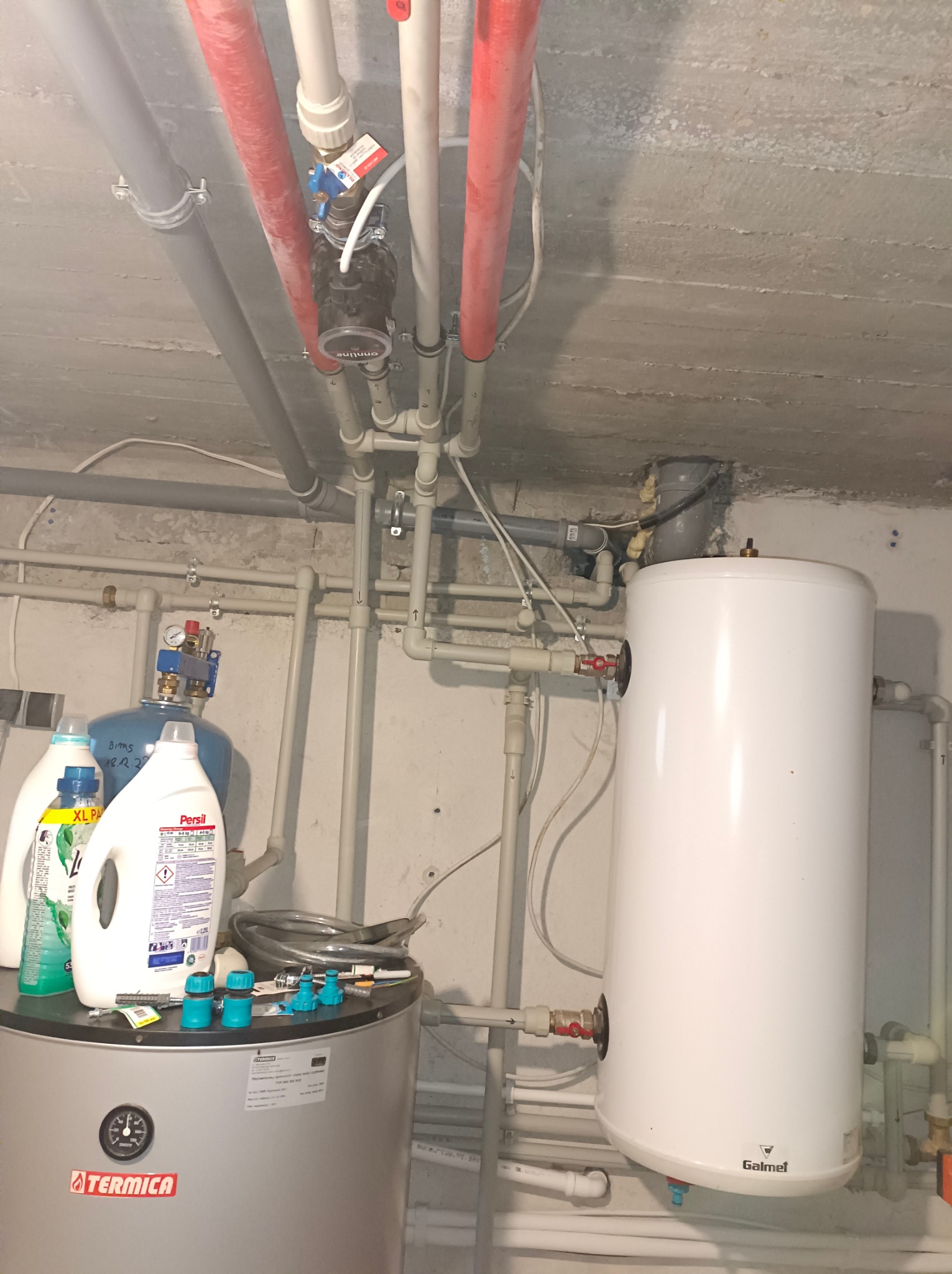 Wnętrze kotłowni z rurami instalacji grzewczej i zbiornikiem na wodę Galmet oraz Termica, widoczne rury miedziane i plastikowe, armatura, manometr, środki czystości i akcesoria hydrauliczne...