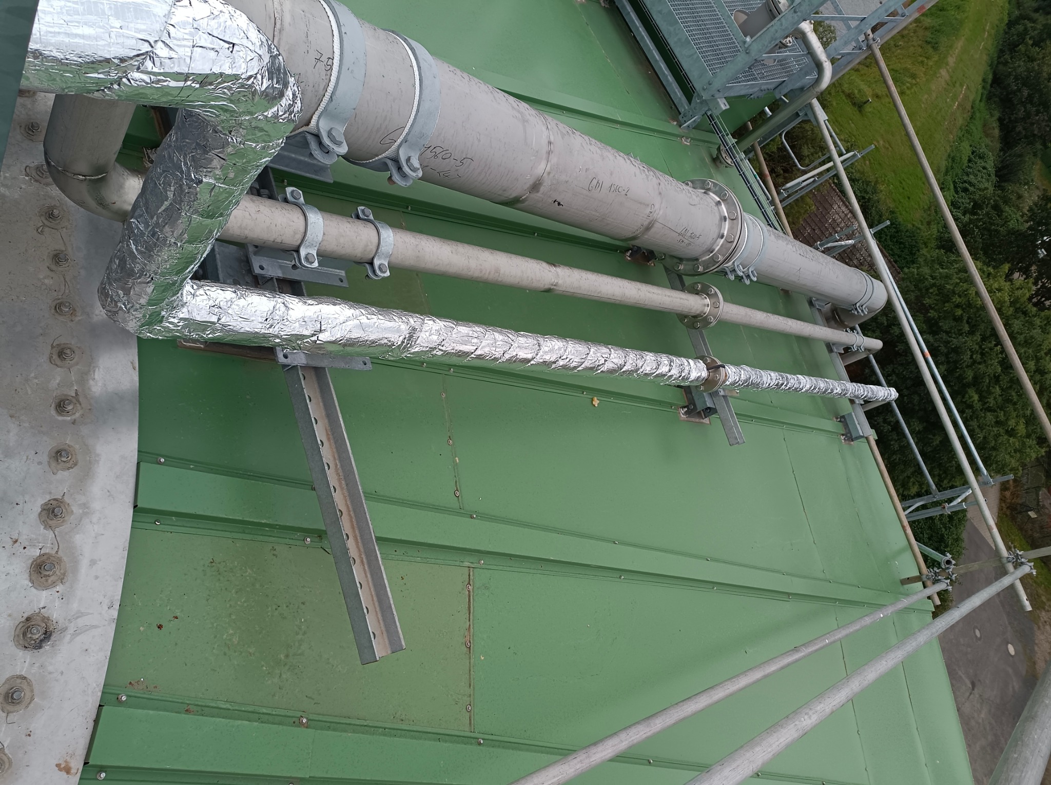 Rurociąg przemysłowy z izolacją termiczną, zamocowany na konstrukcji wsporczej na tle zielonego dachu i elementów rusztowania.