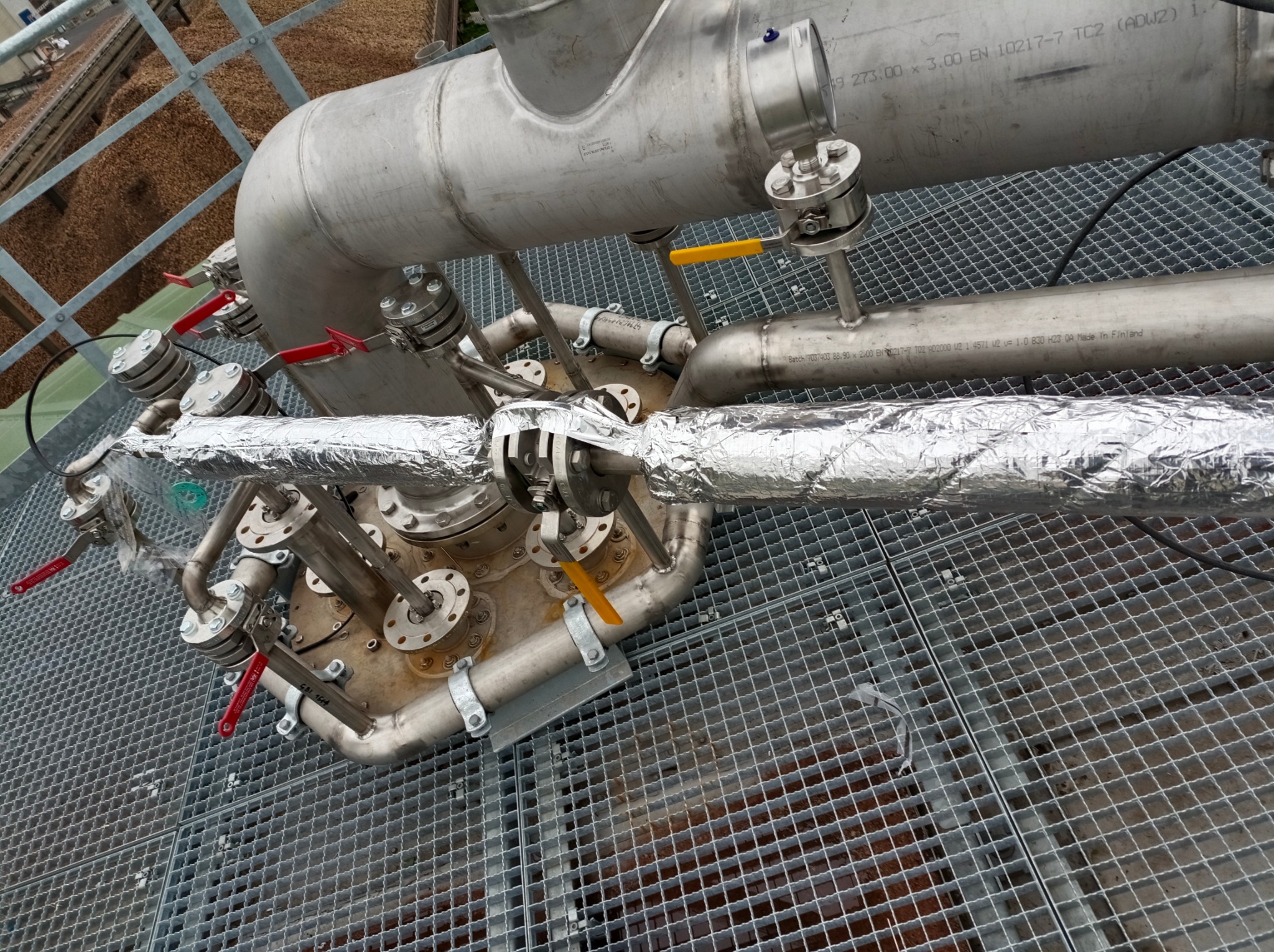 Złożona instalacja przemysłowa z rur i zaworów ze stali nierdzewnej, częściowo owiniętych folią izolacyjną, zamontowana na metalowej kratownicy pod kątem.