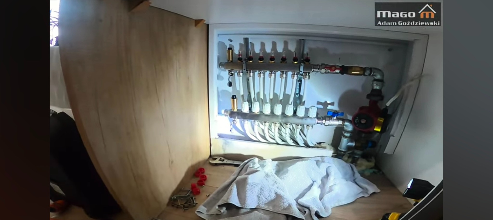 Rozdzielacz ogrzewania podłogowego w obudowie szafki, widoczne rury, zawory, pompa obiegowa, klucz nastawny i akcesoria montażowe.