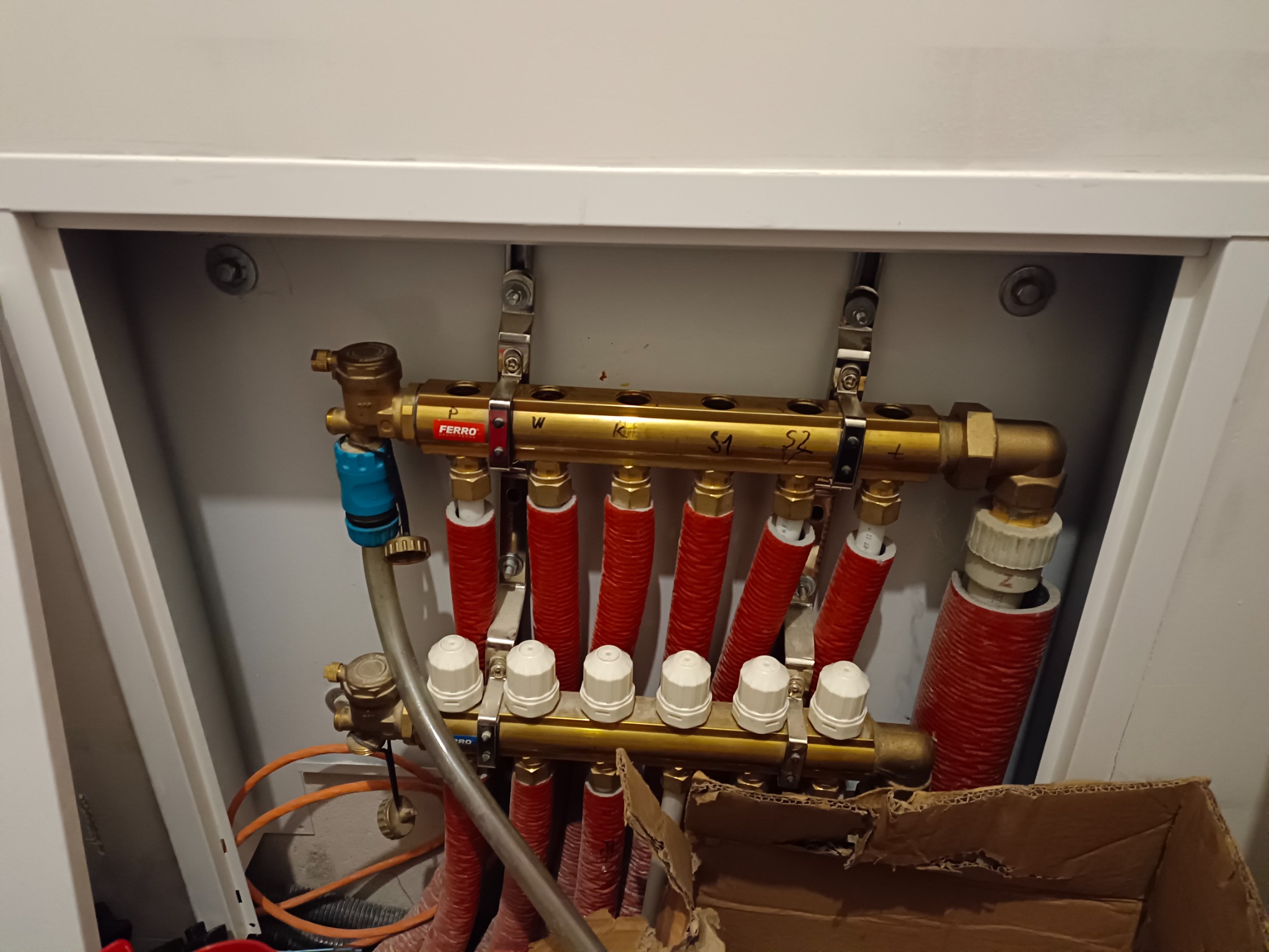 Rozdzielacz instalacji grzewczej podłączony do rur w szafce, widoczne zawory termostatyczne i rury w izolacji termicznej, marka Ferro.