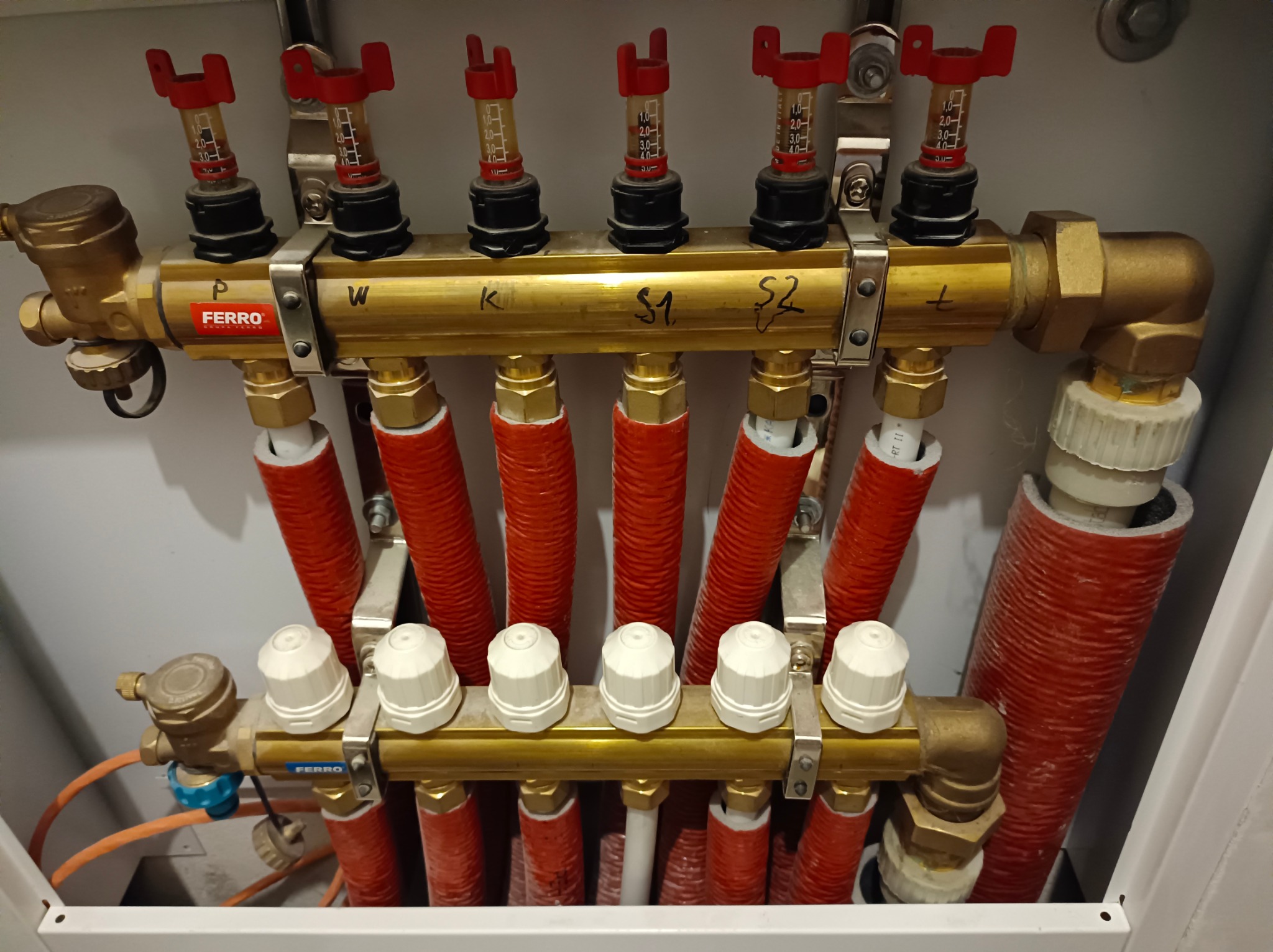 Rozdzielacz instalacji grzewczej marki Ferro, z czerwonymi rurami i przezroczystymi przepływomierzami, zamontowany w szafce, widoczne oznaczenia P, W, K, S1, S2.