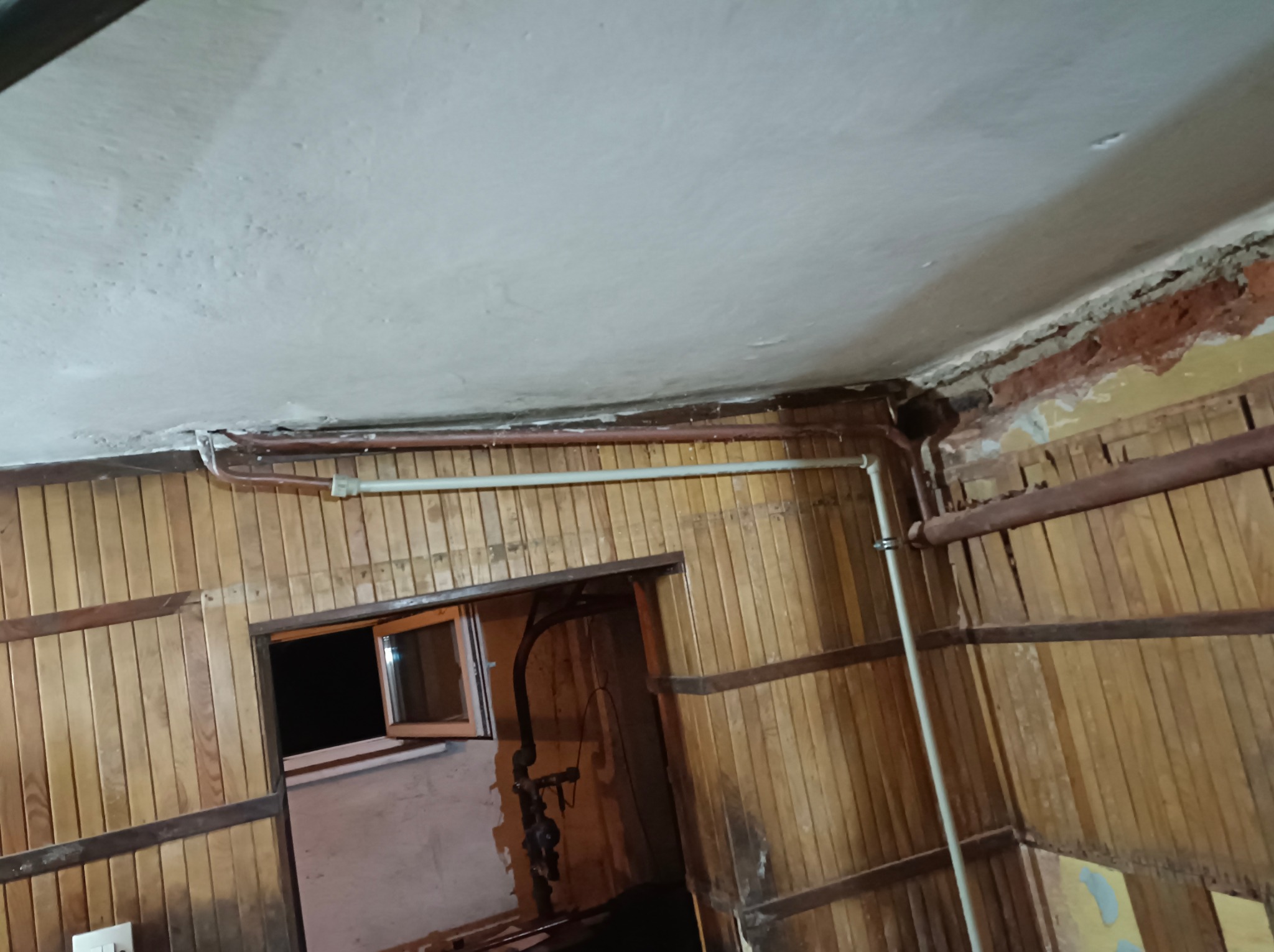 Fragment wnętrza w trakcie remontu, z widocznymi starymi rurami instalacji grzewczej biegnącymi wzdłuż drewnianej boazerii i pod sufitem, odsłonięty fragment muru nad boazerią, małe okno w tle.