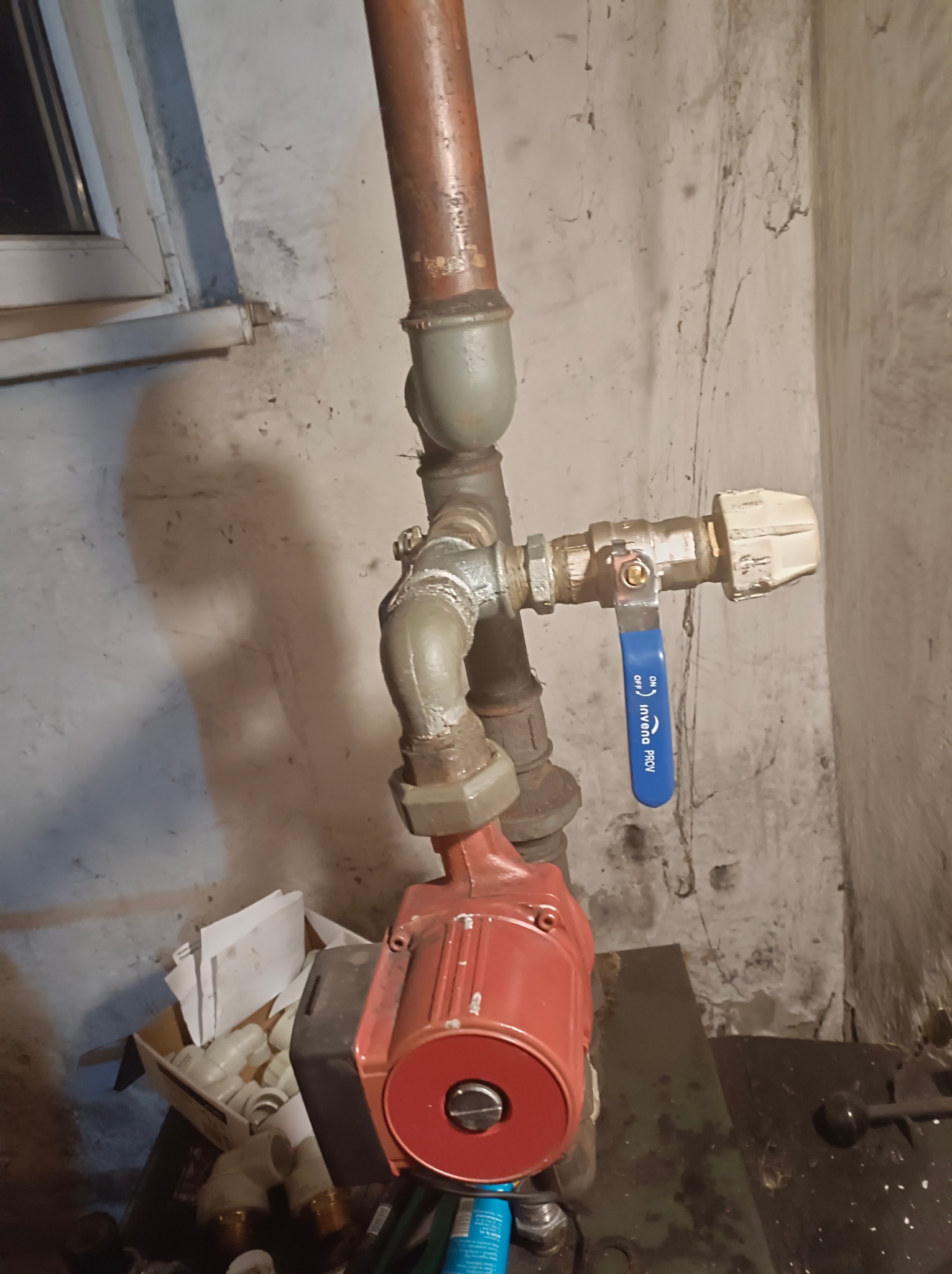 Czerwona pompa obiegowa zamontowana na rurach instalacji grzewczej z zaworem kulowym Invena PROV na tle surowej ściany z widocznymi śladami wilgoci i pajęczynami, obok karton z kształtkami PCV.