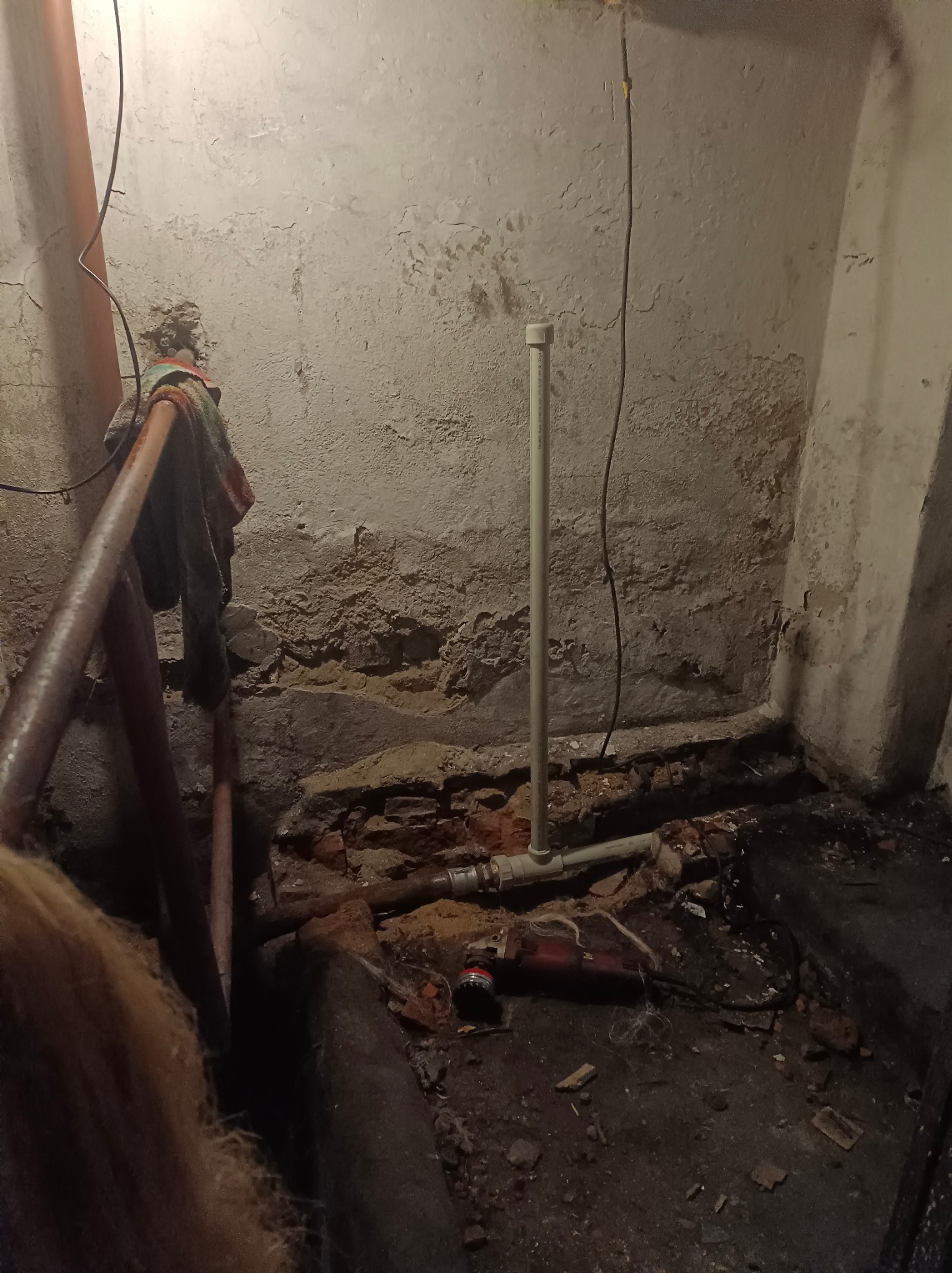 Fragment instalacji hydraulicznej w piwnicy z widoczną rurą PCV, starą ścianą z odpadającym tynkiem i fragmentami gruzu na podłodze, sugerujący remont lub naprawę.