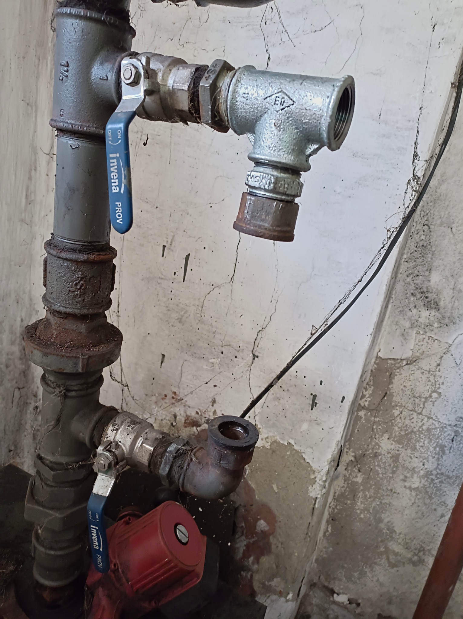 Fragment starej, skorodowanej instalacji hydraulicznej z zaworami Invena, widoczne pajęczyny i ślady po naprawach na ścianie.