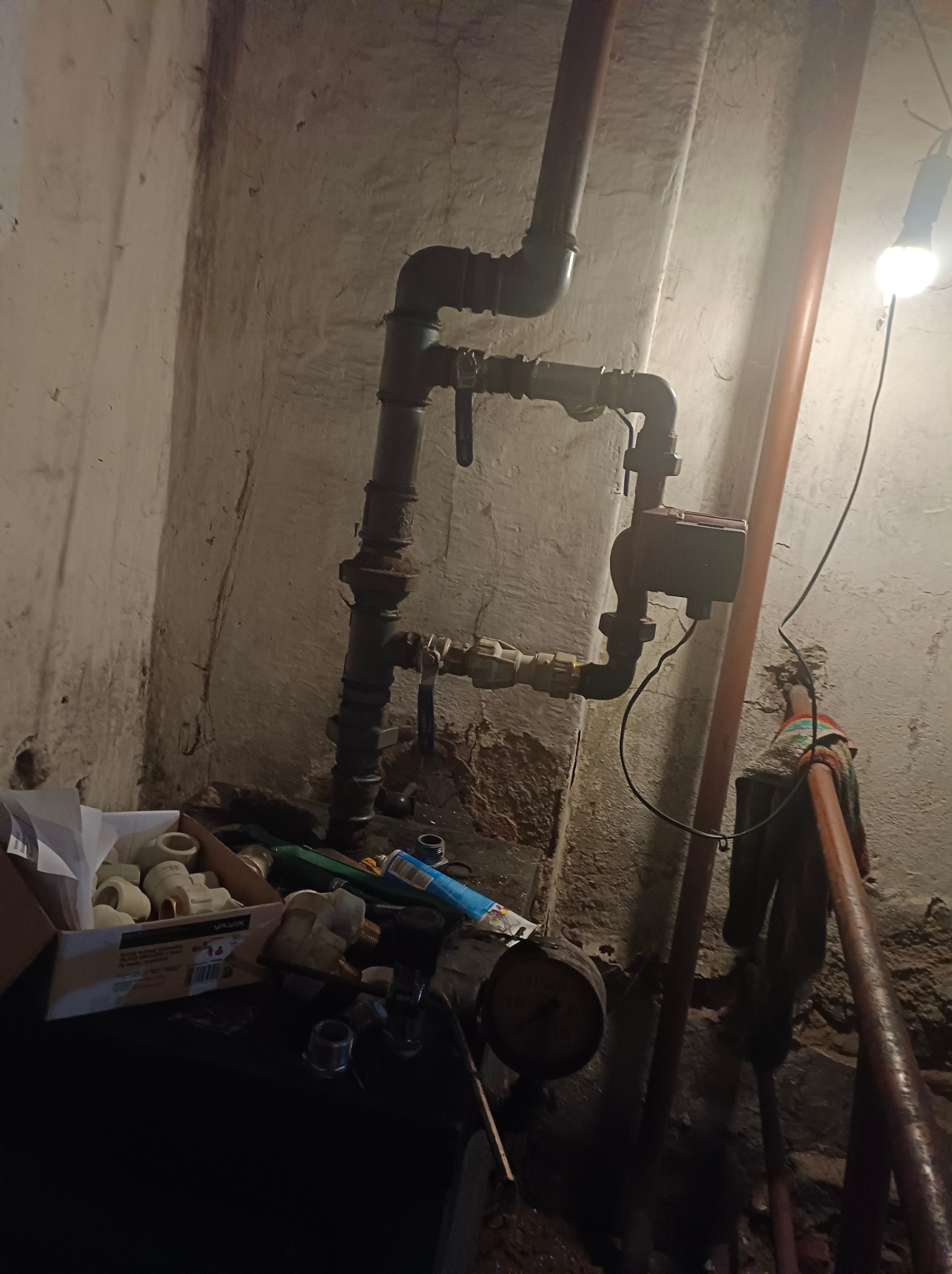 Fragment instalacji grzewczej z pompą obiegową, zaworami i rurami w piwnicy z oświetleniem żarówką, widoczne narzędzia i elementy instalacyjne w pudełku.