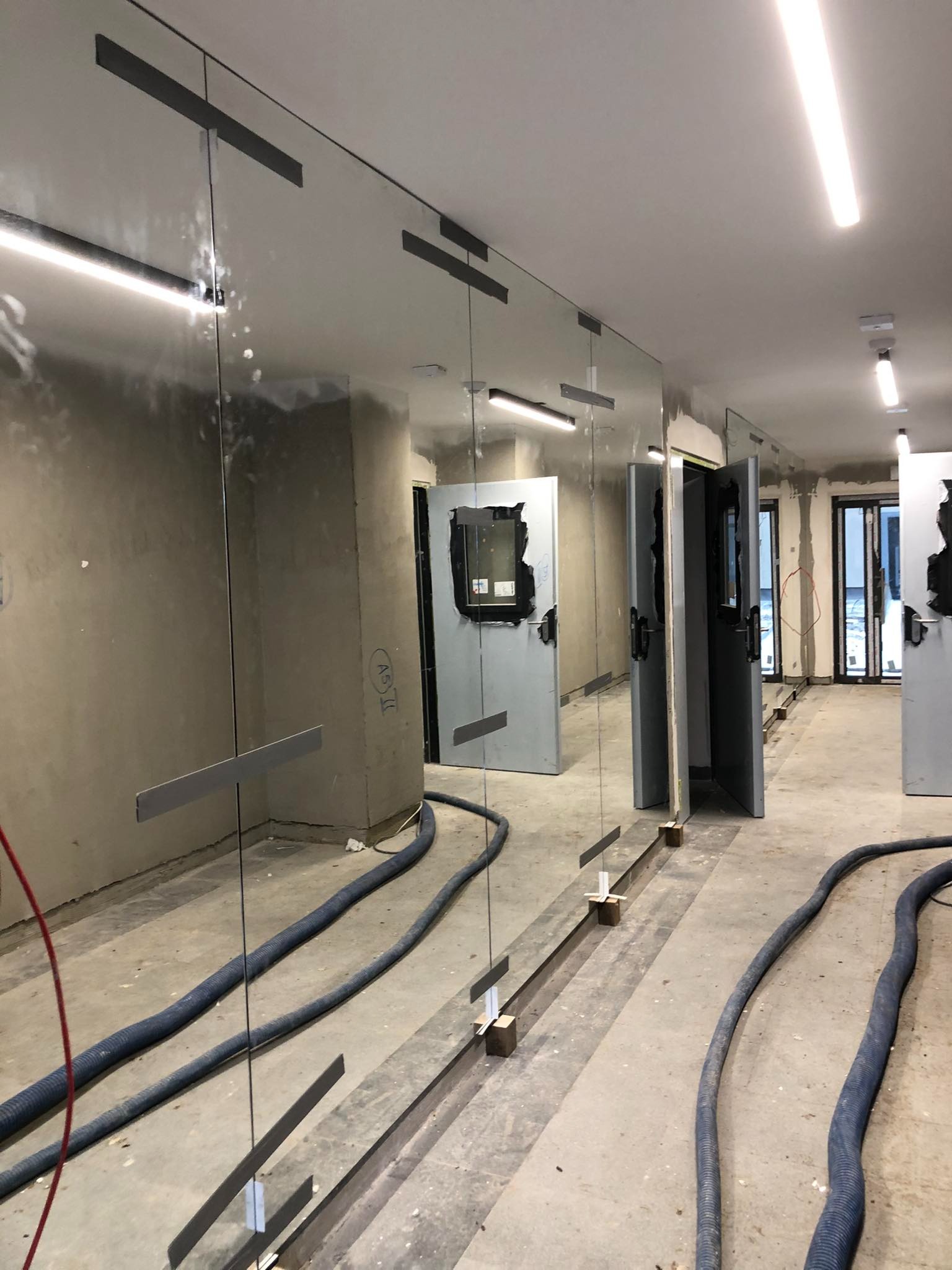 Montaż szklanej ściany działowej z widocznymi metalowymi mocowaniami i tymczasowymi podporami, nieukończone wnętrze z szarymi ścianami i przewodami wentylacyjnymi na podłodze.