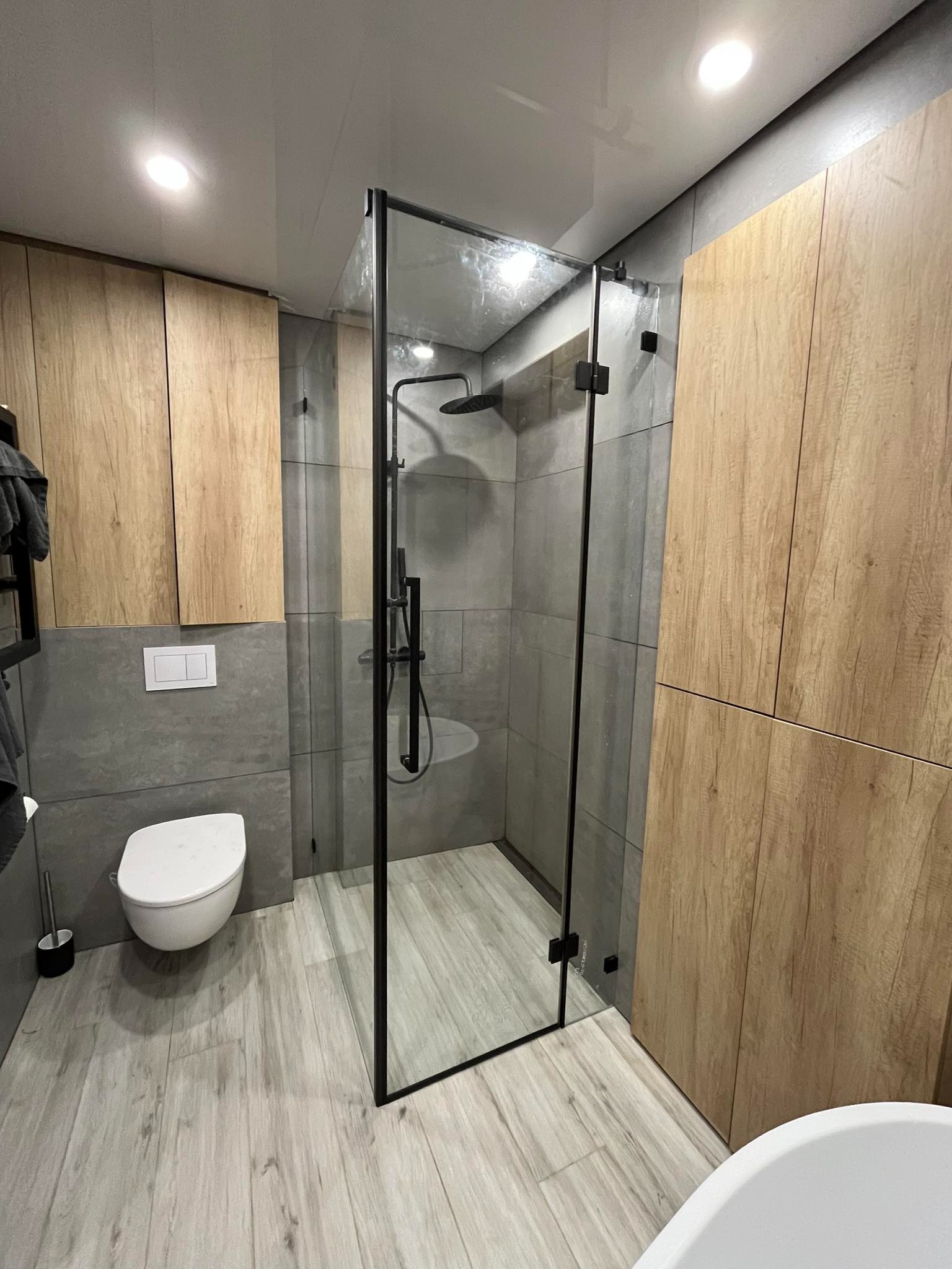 Nowoczesna łazienka z kabiną prysznicową ze szkła hartowanego w czarnej ramie, drewnopodobne szafki wiszące, szare płytki ścienne i podłogowe, biała podwieszana toaleta.
