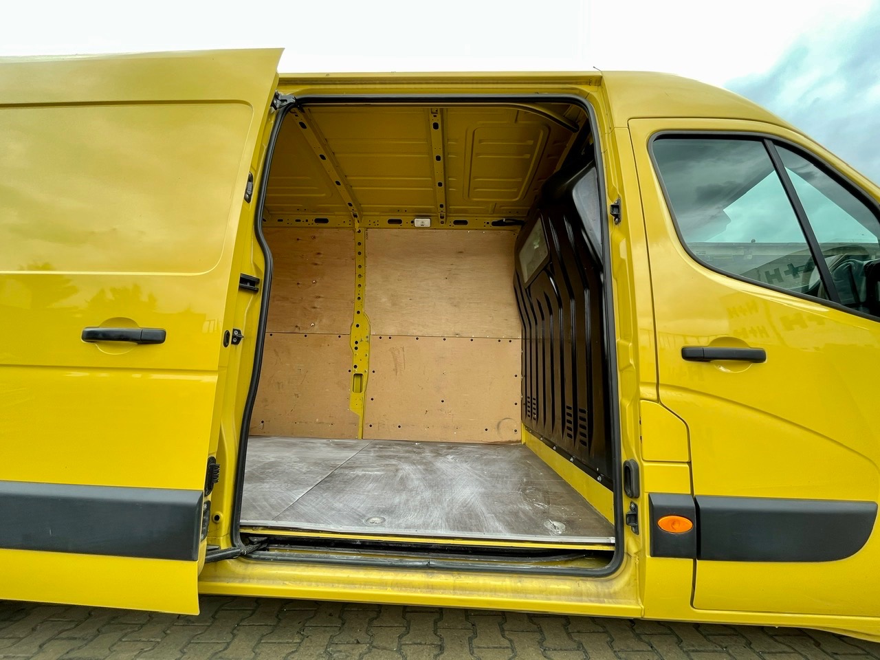 Wnętrze żółtego busa transportowego z otwartymi drzwiami, wyłożone sklejką, przygotowane do przewozu towarów.