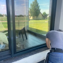 Premium Grodzki - Montaż okna PCV z widokiem na ogród i dom, pracownik w pasie narzędziowym podczas instalacji, widoczne zabezpieczenia przeciwwilgociowe.