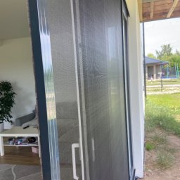 Premium Grodzki - Szara moskitiera przesuwna na oknie, widok z wnętrza domu na zewnątrz, z widocznym ogrodem i domem w tle.