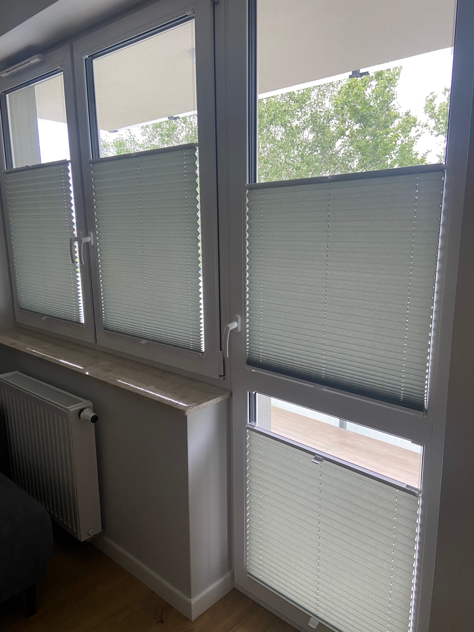 Białe okna PCV z zamontowanymi plisowanymi roletami wewnętrznymi, widok z wnętrza pomieszczenia na zewnątrz, częściowe zasłonięcie okien, radiator pod parapetem.