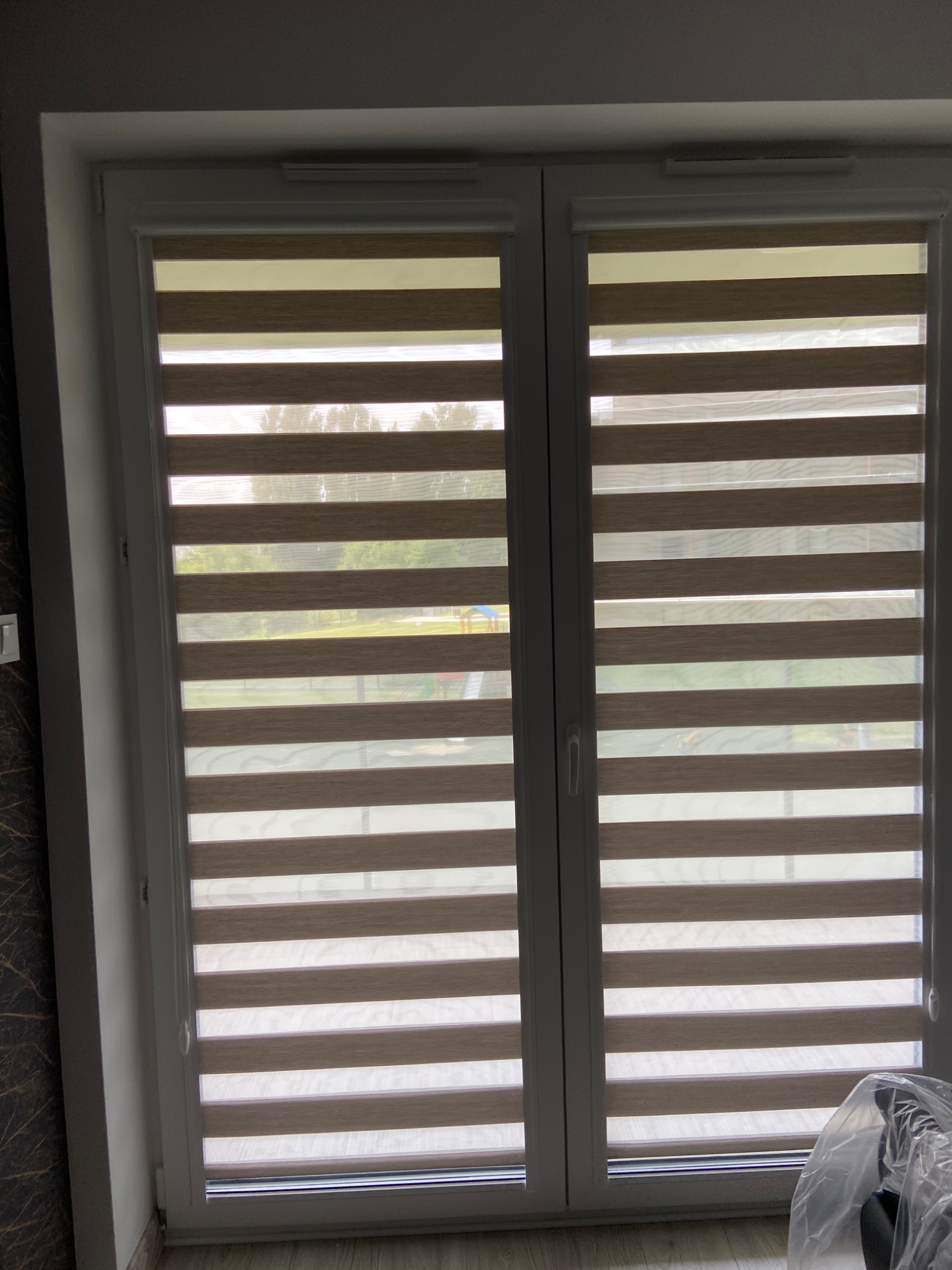 Dwuskrzydłowe okno z zamontowanymi roletami dzień i noc w odcieniach beżu i bieli, widok na zewnątrz z placem zabaw w tle.