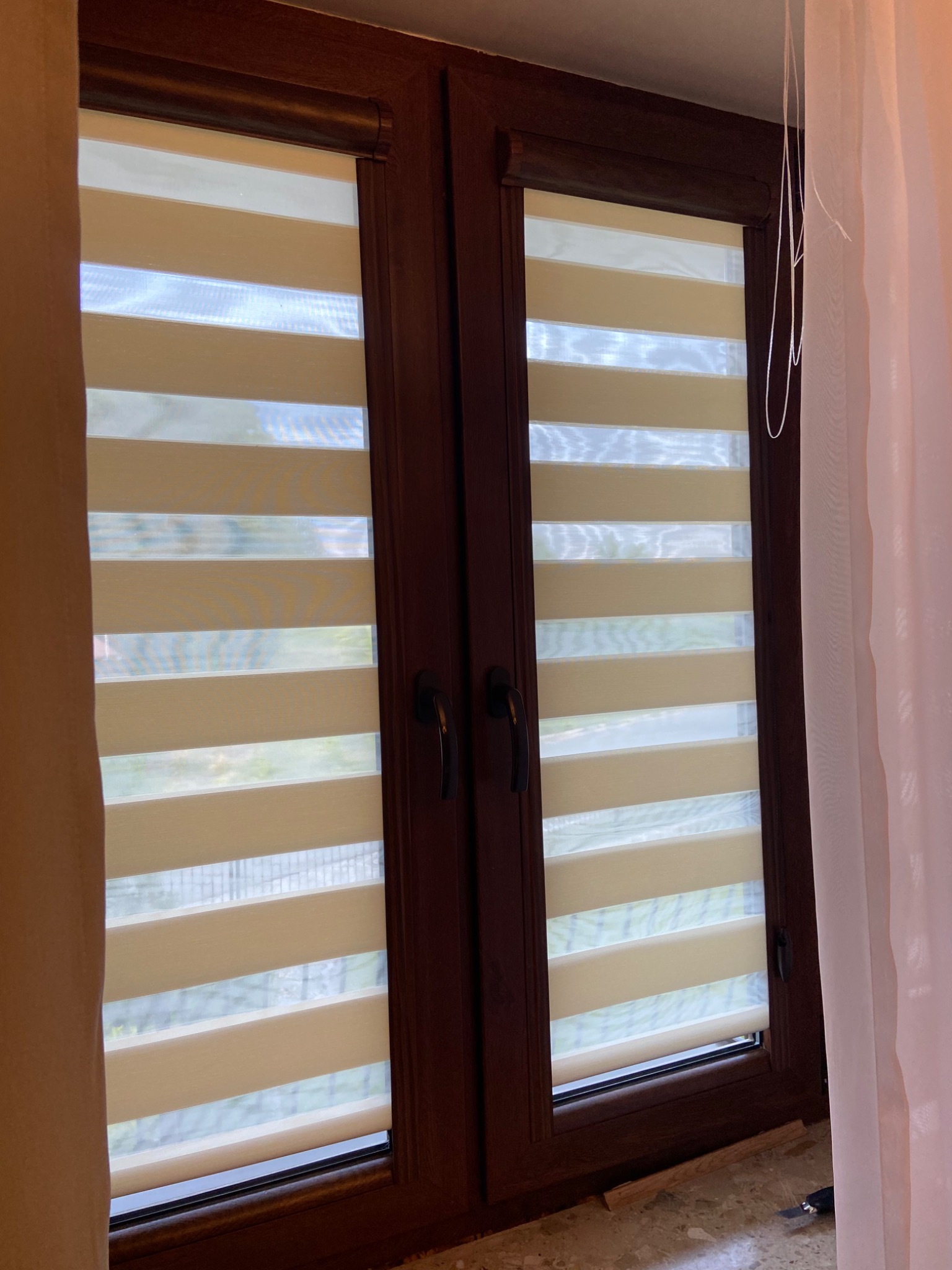 Drewniane okno z zamontowaną roletą wewnętrzną typu dzień i noc w beżowo-białe pasy, zasłonięte częściowo białym i beżowym materiałem.