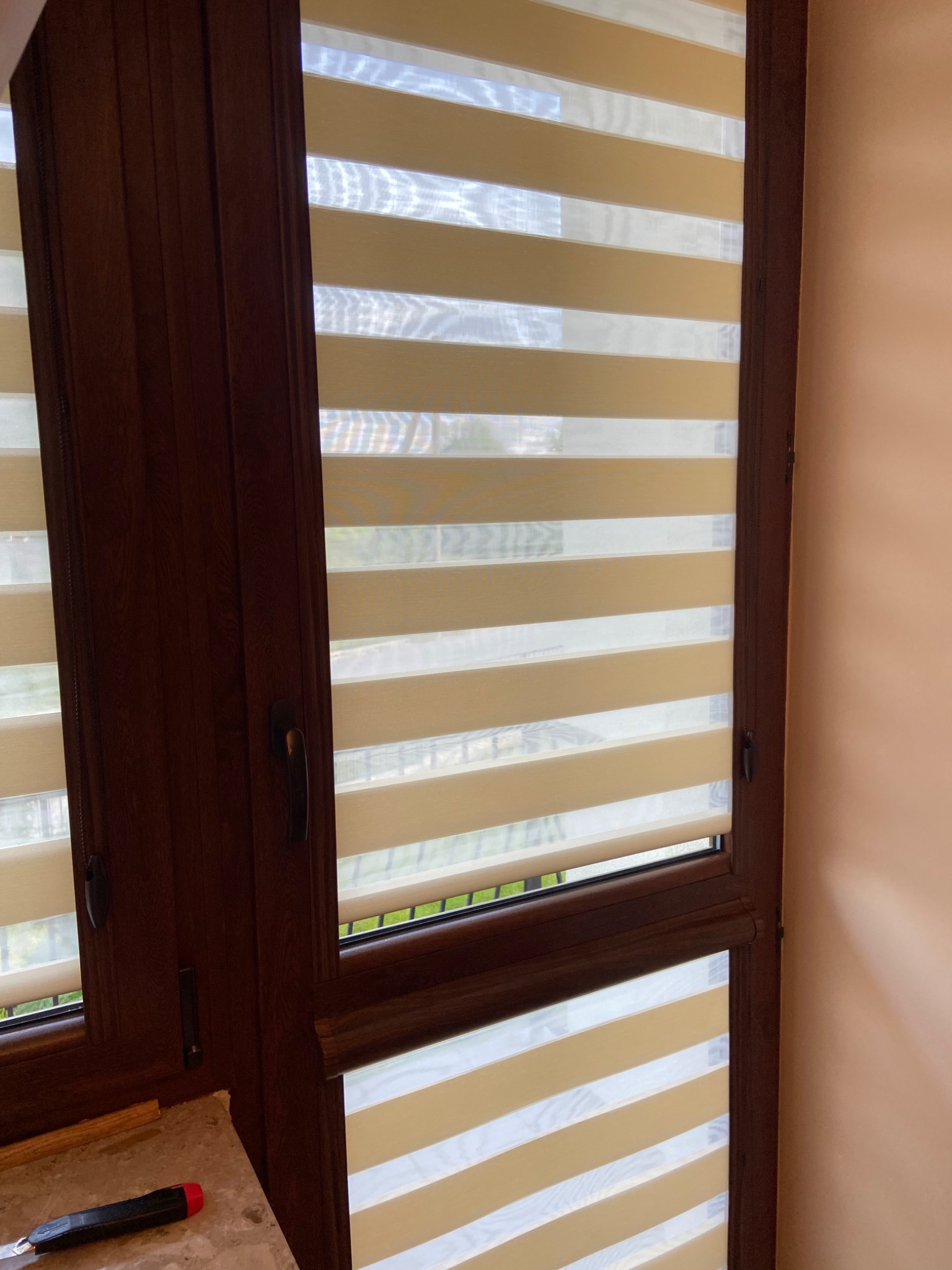 Okno z brązową ramą i zamontowanymi roletami dzień-noc w beżowo-białe pasy, widok z bliska, na parapecie leży nóż do tapet.