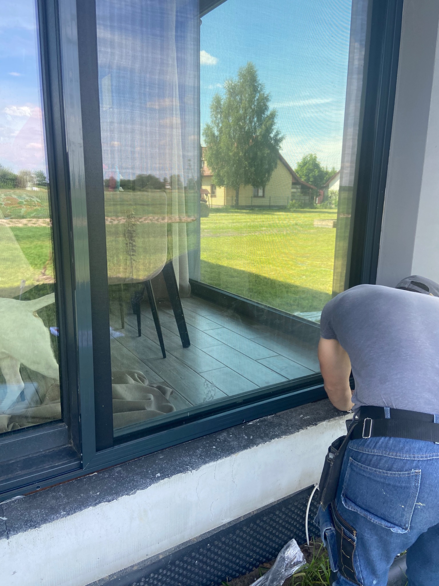 Montaż okna PCV z widokiem na ogród i dom, pracownik w pasie narzędziowym podczas instalacji, widoczne zabezpieczenia przeciwwilgociowe.