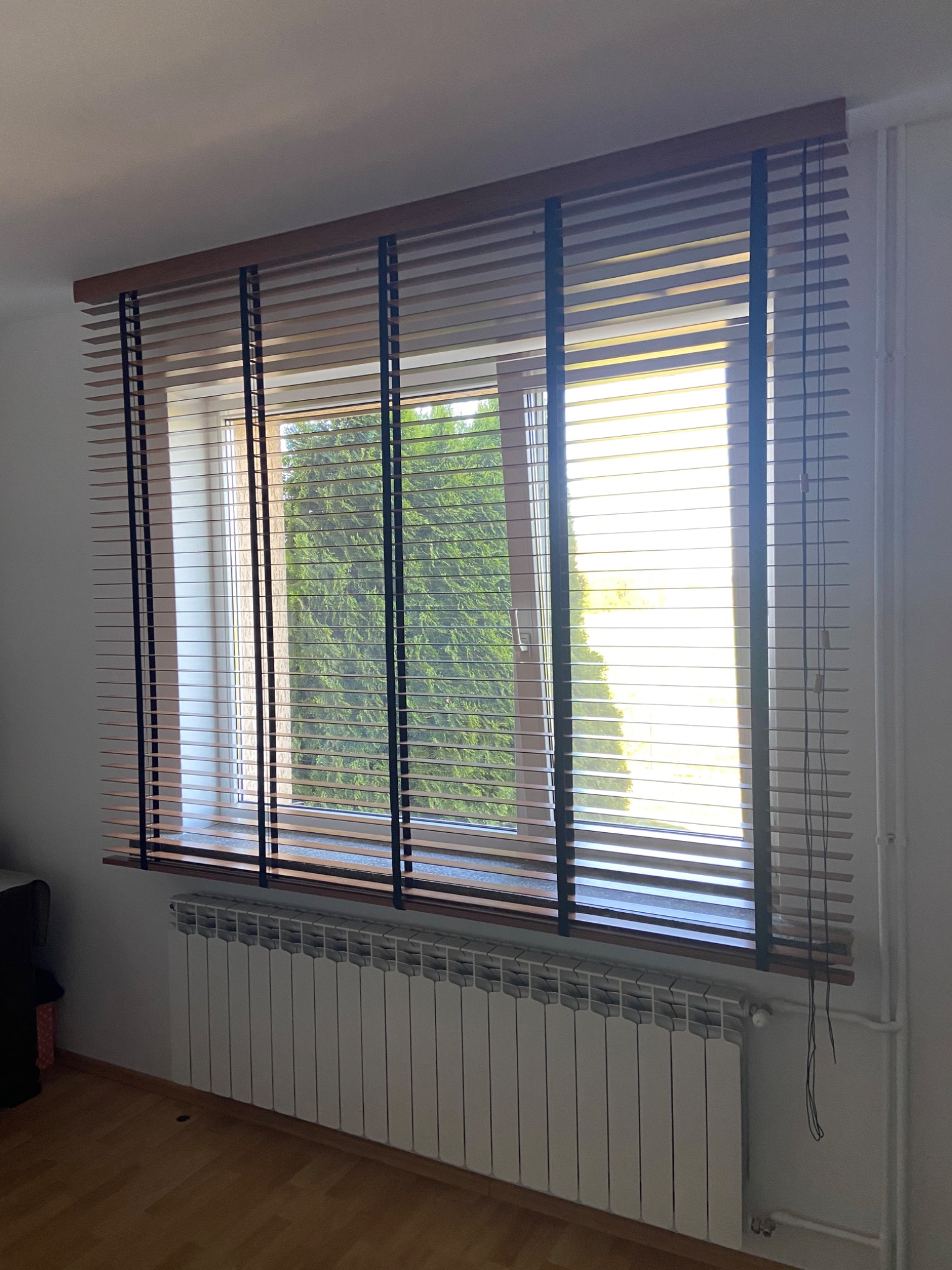 Zamknięte drewniane żaluzje poziome w oknie, widok na zieleń za oknem, biały grzejnik pod oknem.