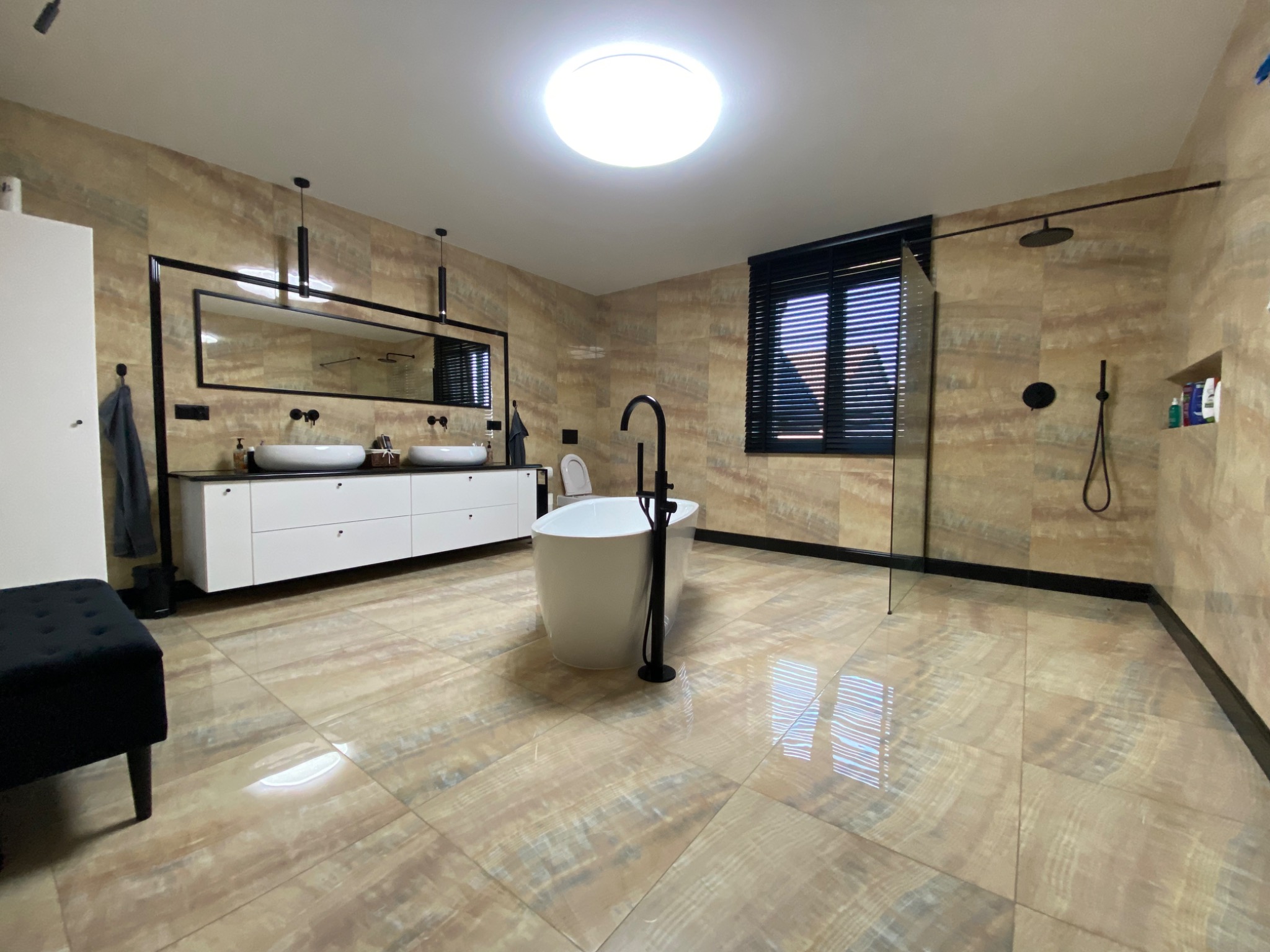 Nowoczesna łazienka z wolnostojącą wanną, podwójną umywalką na białej szafce, czarną armaturą i żaluzjami w oknie.