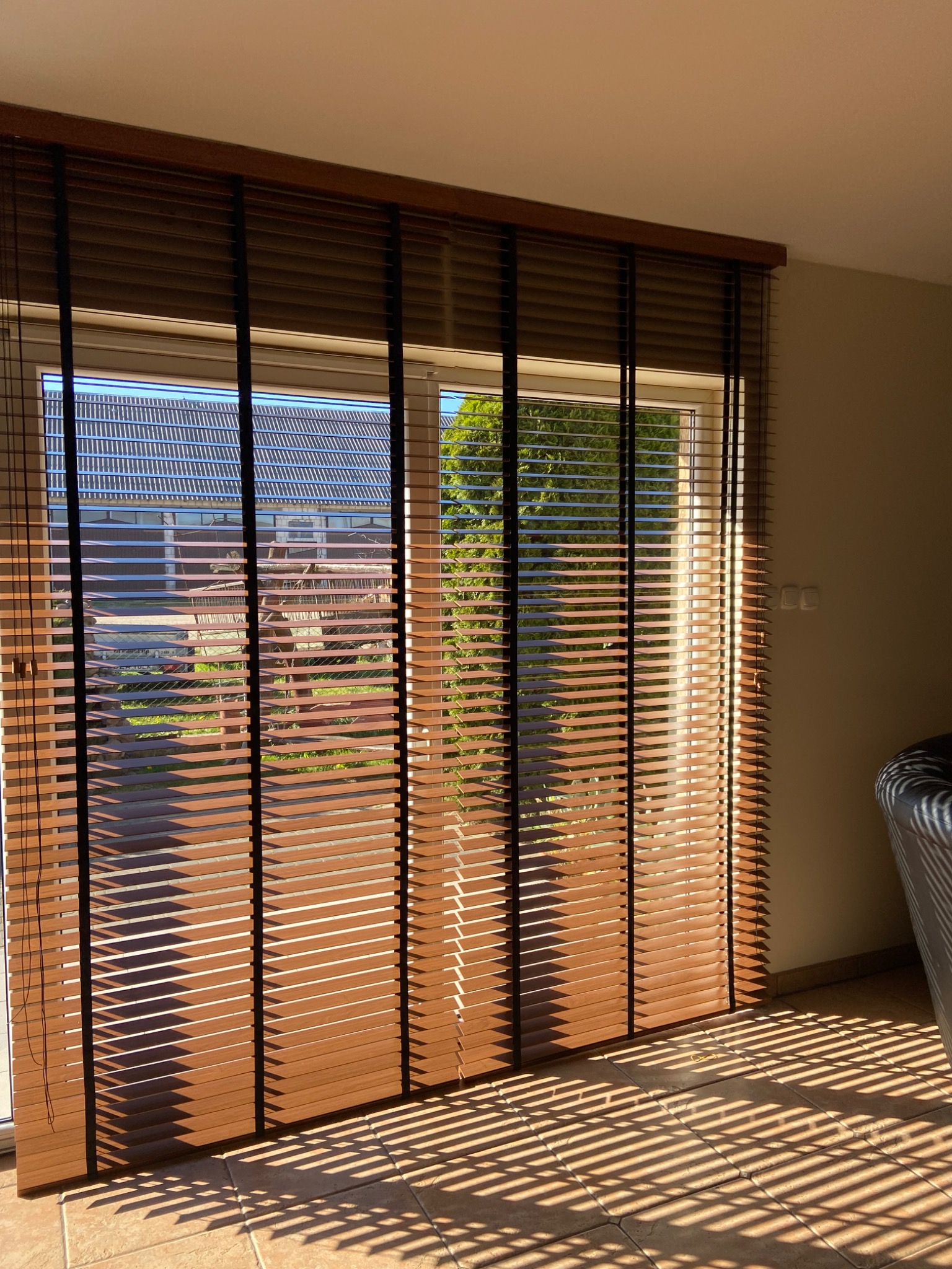 Brązowe żaluzje poziome z drewnianymi lamelami i czarnymi taśmami, częściowo zasłonięte okno z widokiem na ogród i dom sąsiada, promienie słońca tworzą wzór na podłodze.