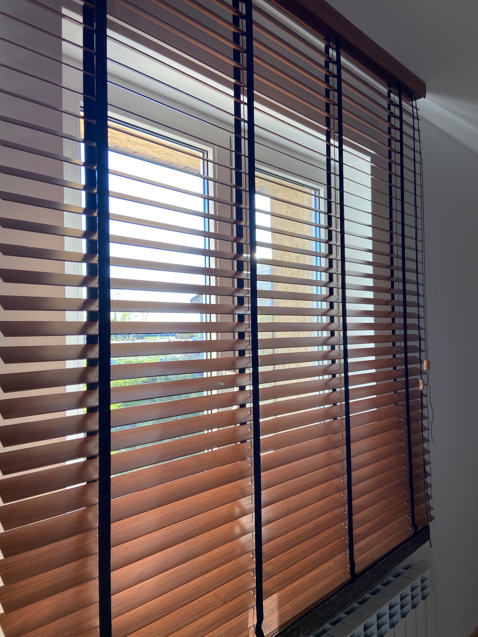 Drewniane żaluzje poziome z czarnymi sznurkami na oknie PCV, częściowo zasłonięte, widok na zieleń za oknem.