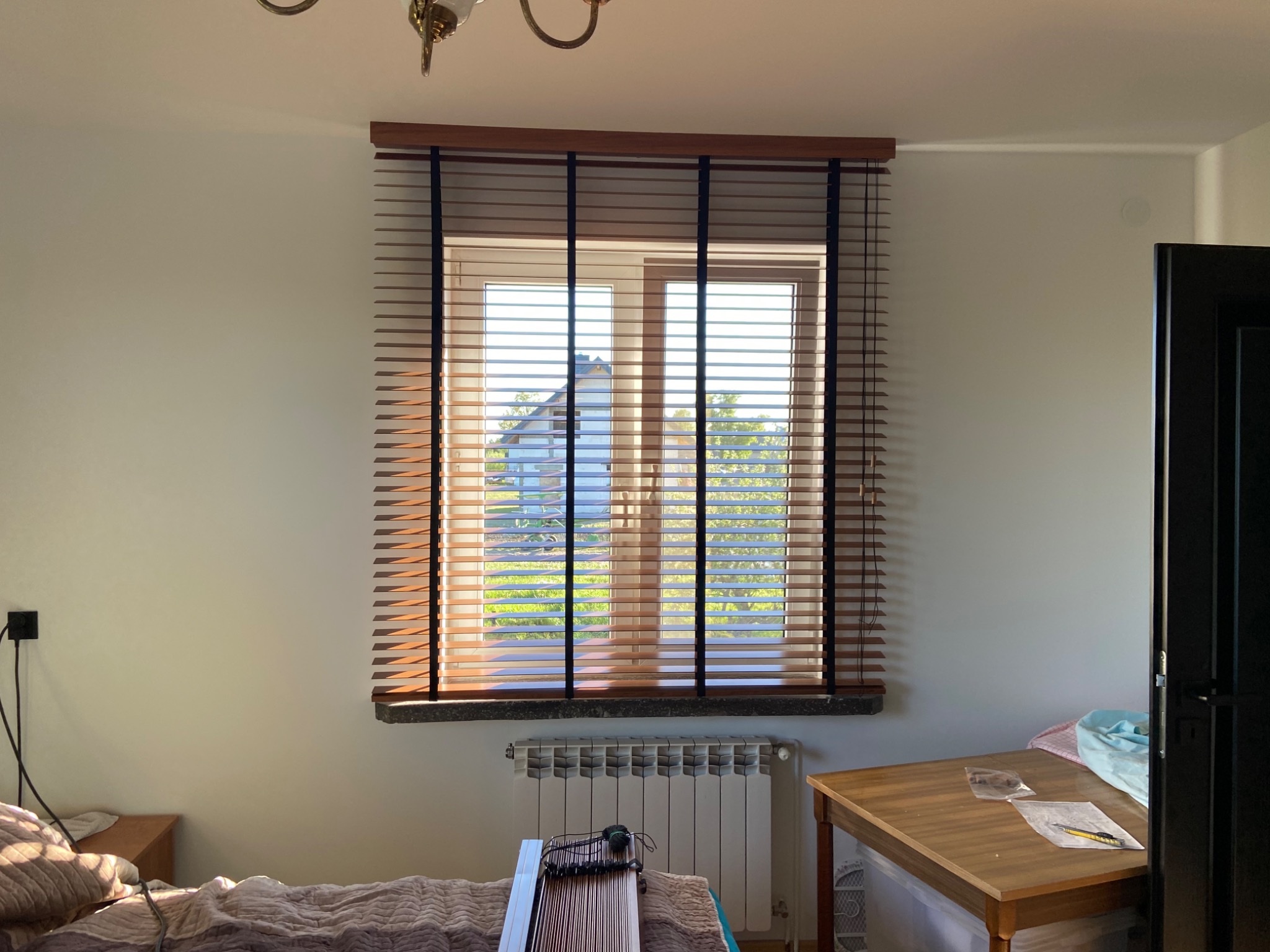 Zamknięte drewniane żaluzje poziome z czarnymi taśmami na oknie w pokoju, widok na zielony ogród na zewnątrz.