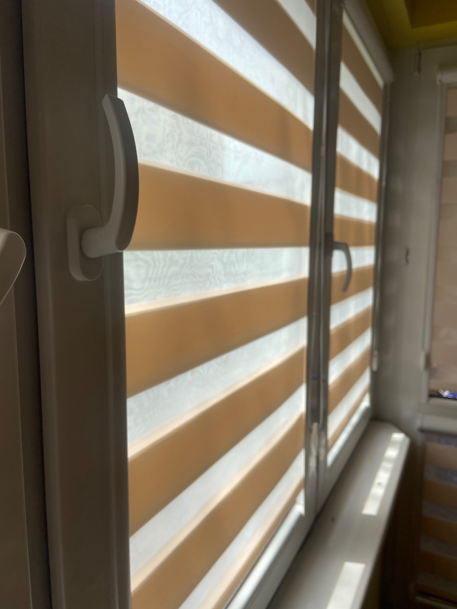 Detal okna z białymi ramami i poziomymi żaluzjami w odcieniach beżu i bieli, z widocznym uchwytem okiennym.