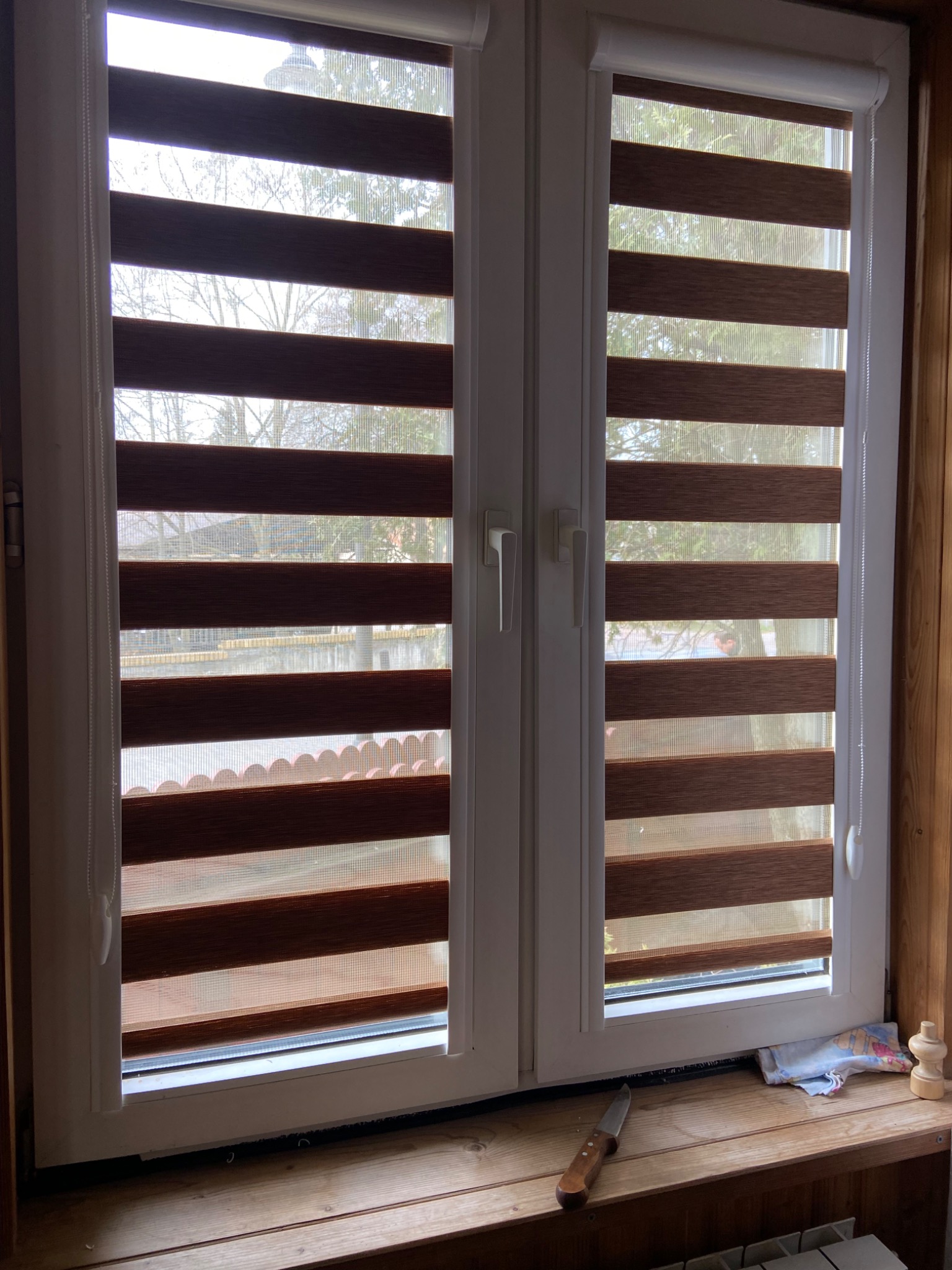 Białe okno z brązowo-białymi roletami dzień-noc, na parapecie nóż i szmatka, widok na drzewa za oknem.