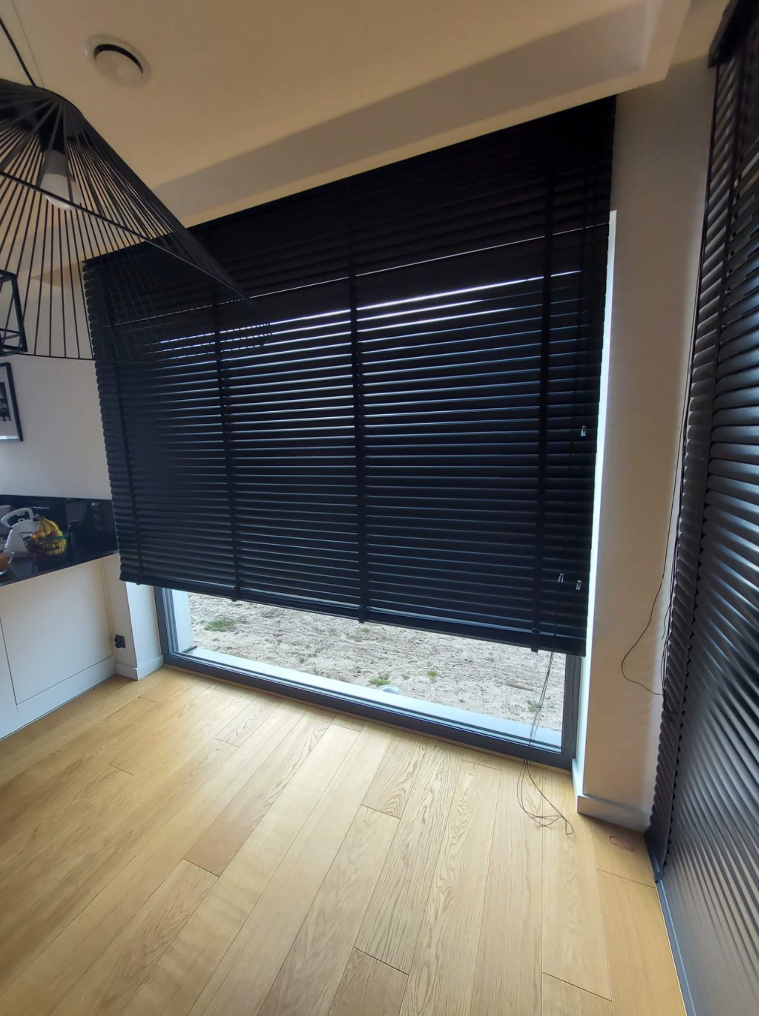 Czarne żaluzje poziome zamontowane w oknie, widok z wnętrza pomieszczenia z parkietem, fragment czarnej lampy wiszącej w rogu kadru.