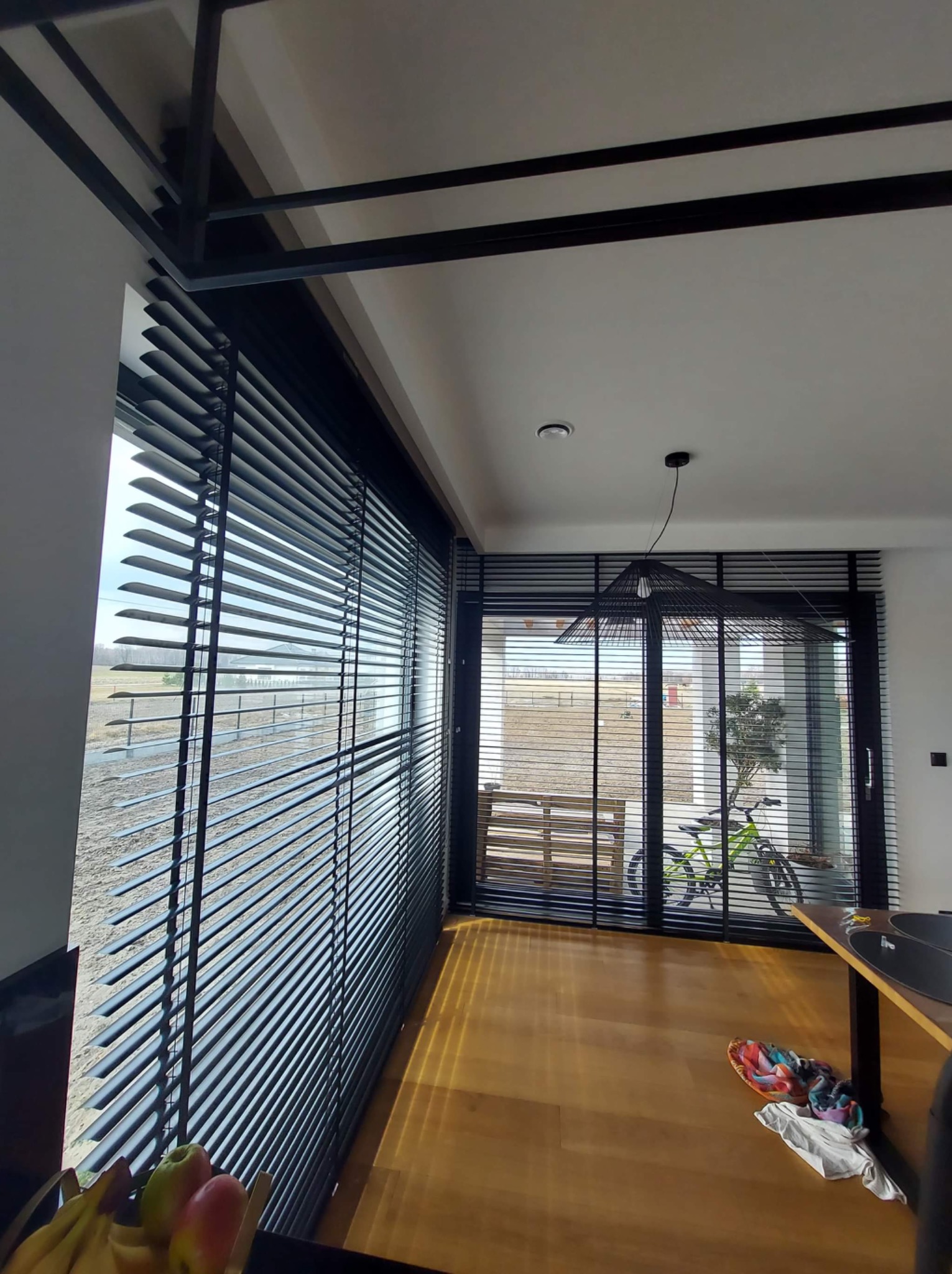 Wnętrze z dużymi oknami przesłoniętymi ciemnymi żaluzjami aluminiowymi, przez które wpada światło słoneczne; na zewnątrz widoczny krajobraz.
