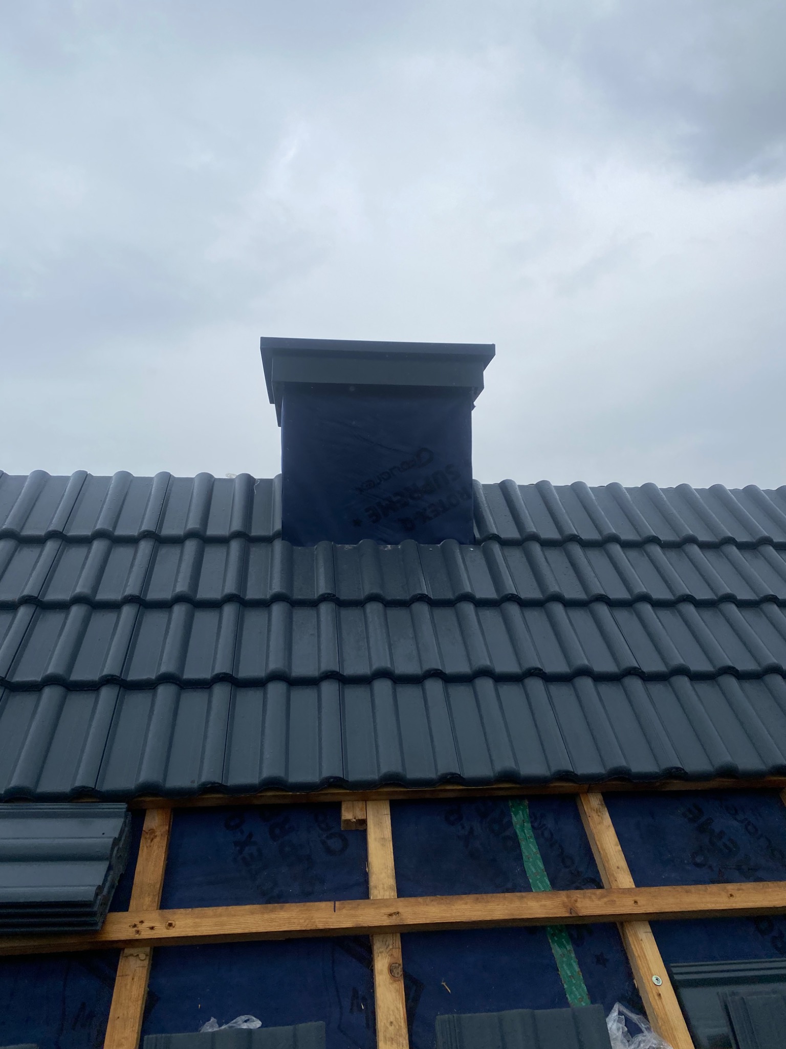 Ciemna dachówka ceramiczna na dachu w trakcie montażu, widoczny komin obłożony membraną oraz drewniana konstrukcja łatowania pod dachówką.