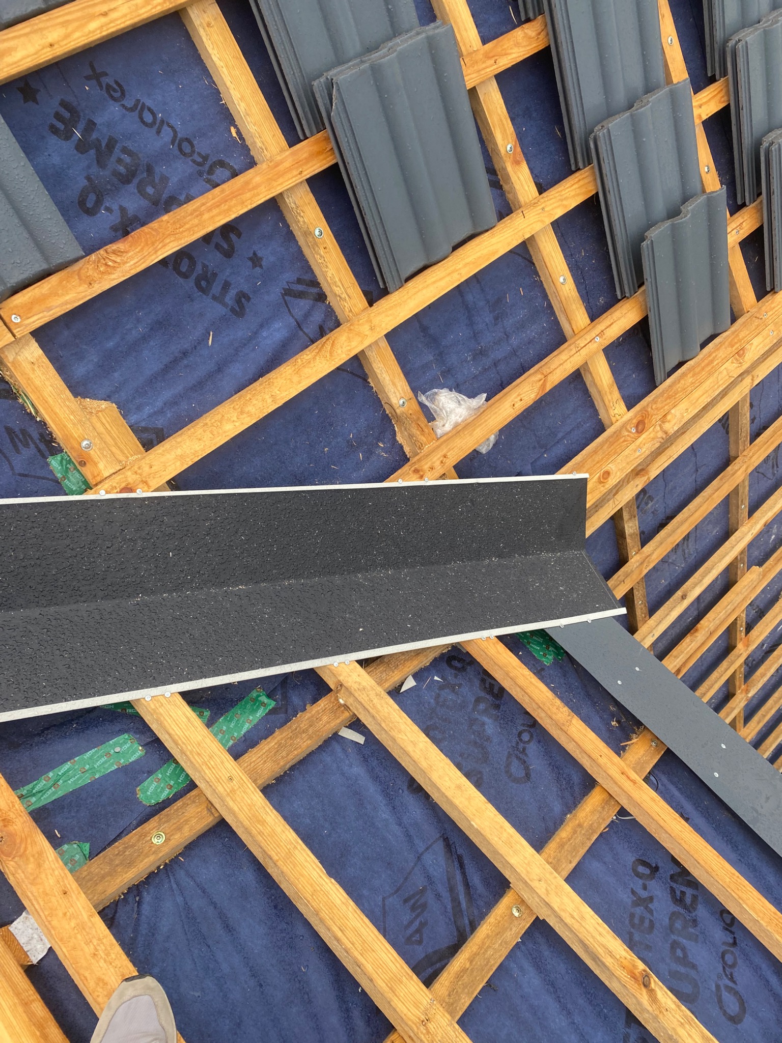 Widok z góry na konstrukcję drewnianą dachu z niebieską membraną, częściowo pokrytą szarymi dachówkami i elementami obróbki blacharskiej, w trakcie montażu.