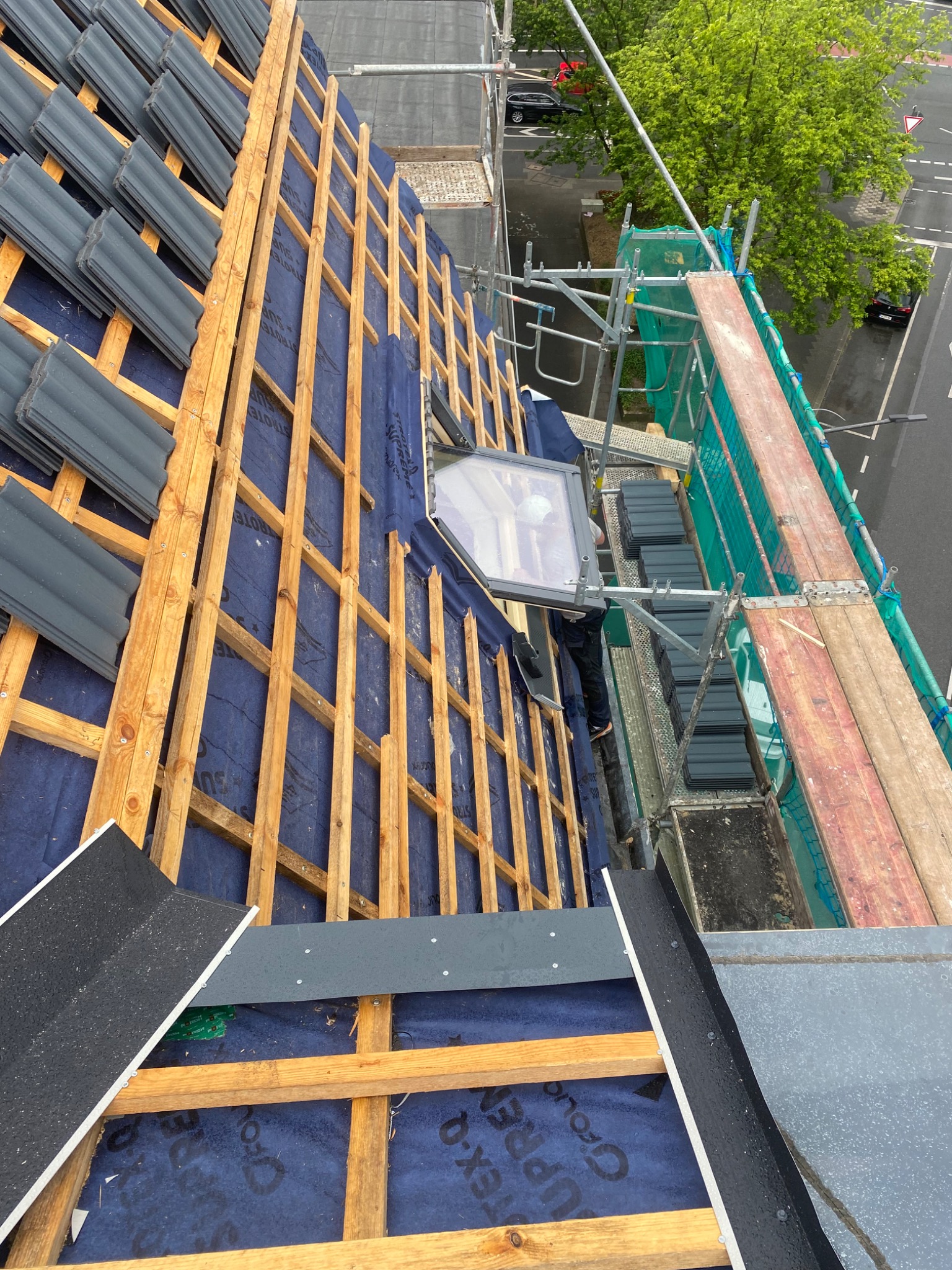 Montaż okna dachowego na drewnianej konstrukcji dachu pokrytej niebieską membraną, widoczne częściowo ułożone szare dachówki i rusztowanie z boku.