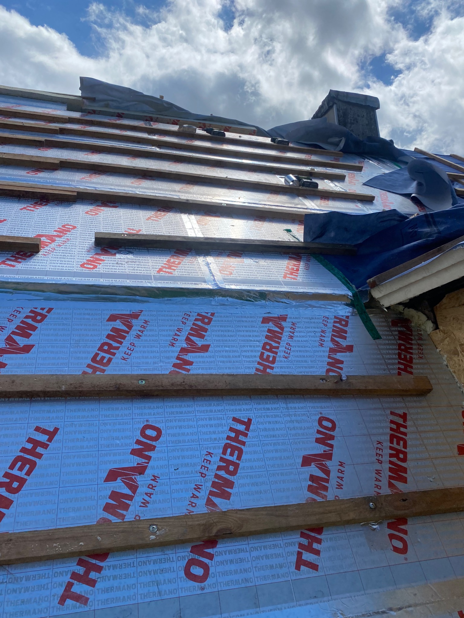 Montaż izolacji termicznej dachu z widocznymi łatami i folią termoizolacyjną z nadrukiem producenta, widok na komin i niebo.